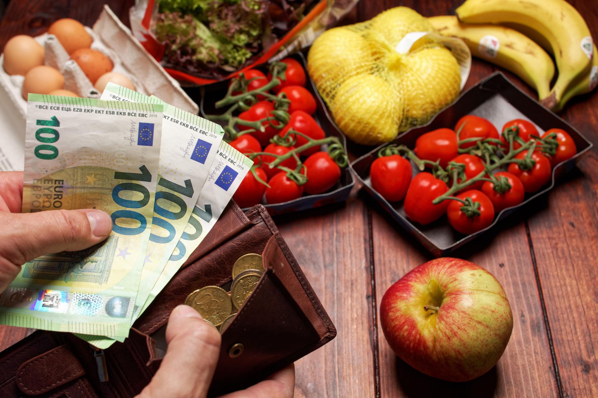 EIne Hand hält dre einhundert Euro-Scheine die andere Hand hält einen Geldbeutel im Hintergund liegt ein Einkauf mit Obst Gemüse und Eiern.