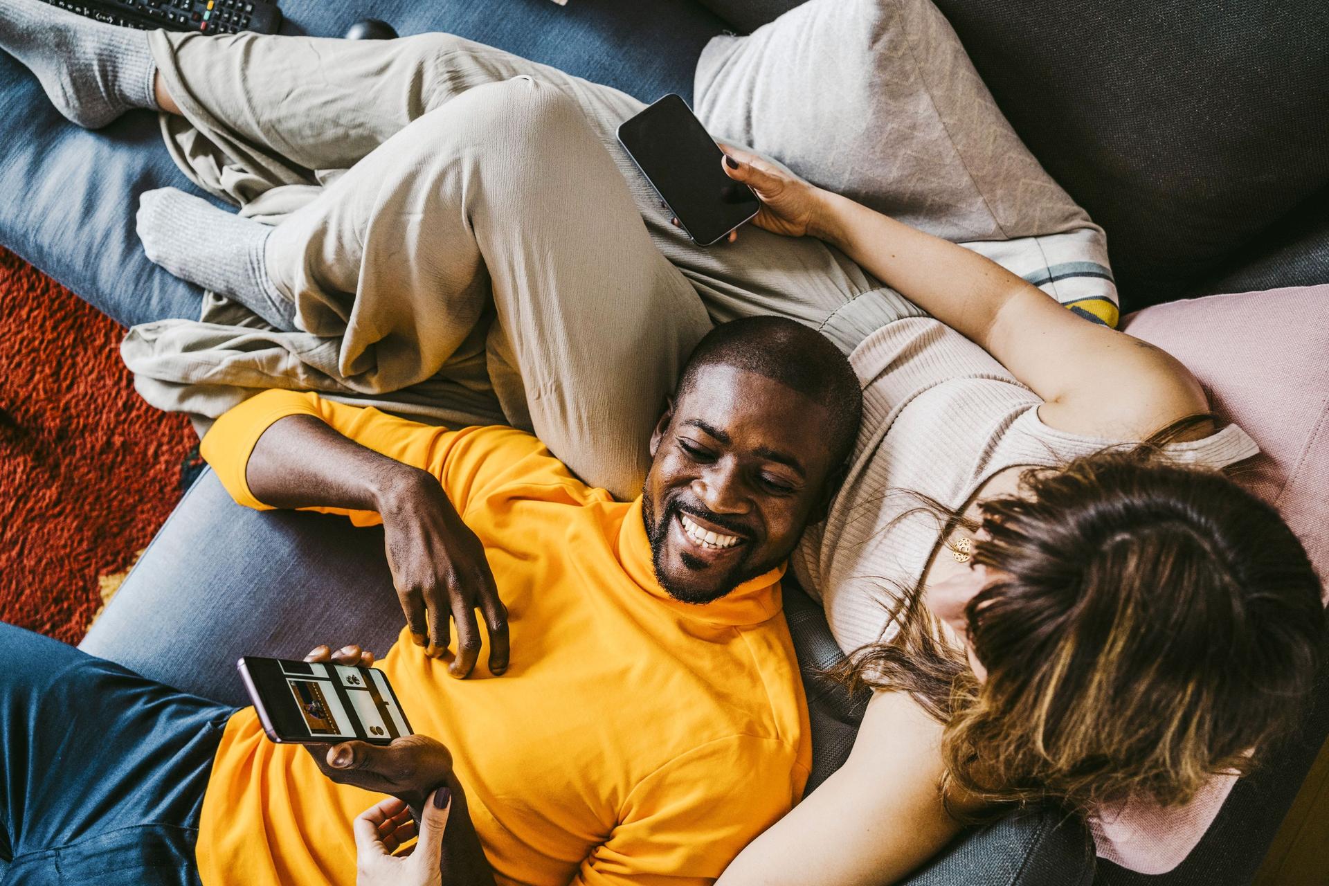 Lachender Mann mit Smartphone in der Hand, der mit seiner Freundin auf dem Sofa liegt und ihr etwas auf dem Bildschirm zeigt.