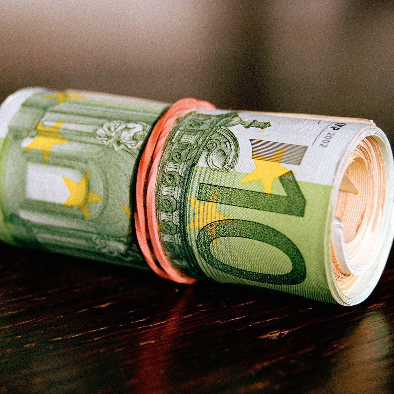 Ein mit einem Gummiband zusammengerolltes Bündel von verschiedenen Geldscheinen liegt auf einer reflektierenden Oberfläche.
