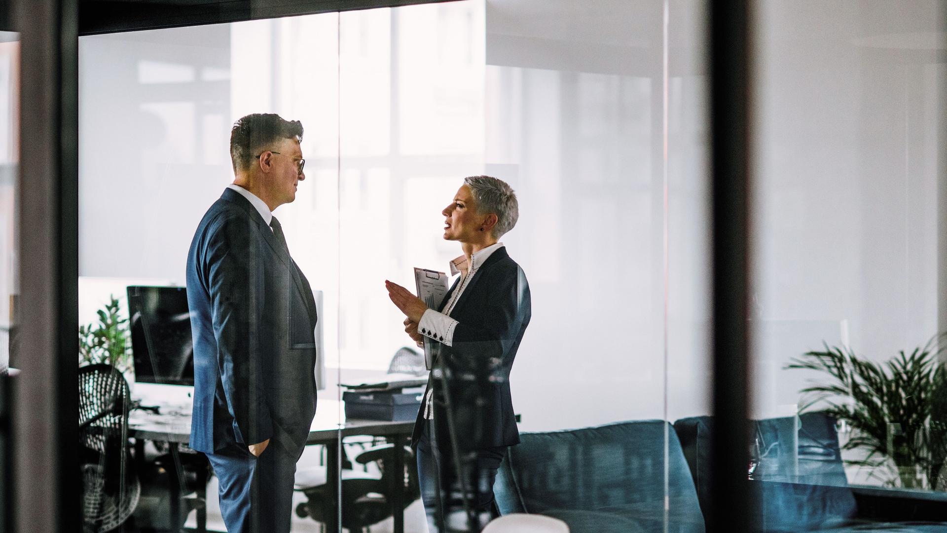 Ein Mann und eine Frau in Business-Kleidung sprechen miteinander  hinter einer Glasscheibe in einem modernen Büro.