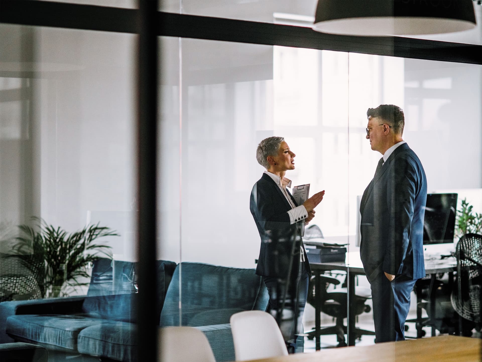 Ein Mann und eine Frau in Business-Kleidung sprechen miteinander  hinter einer Glasscheibe in einem modernen Büro.