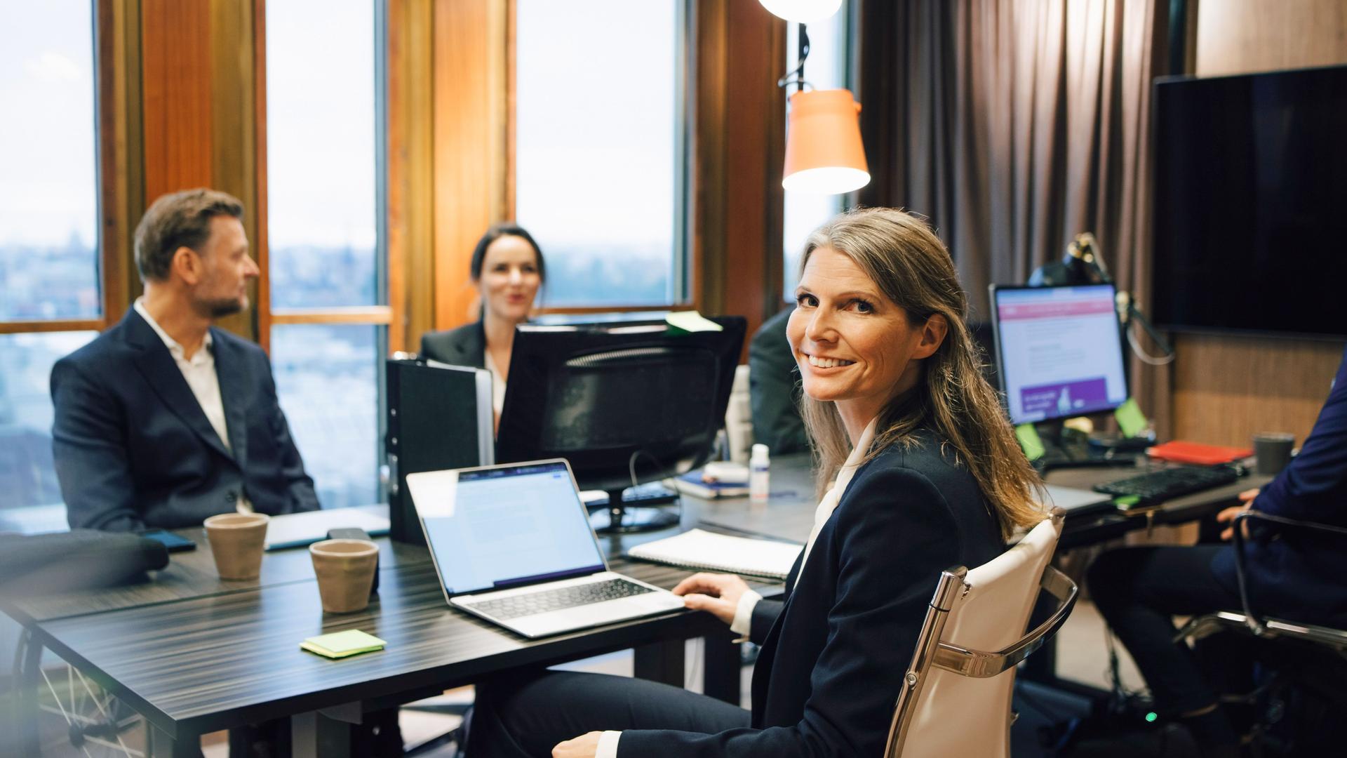 Eine Frau in Business-Kleidung dreht sich während eines Meetings mit drei Männern und einer Frau in einem Konferenzraum lächelnd in die Kamera. Sie hat einen aufgeklappten Laptop vor sich.