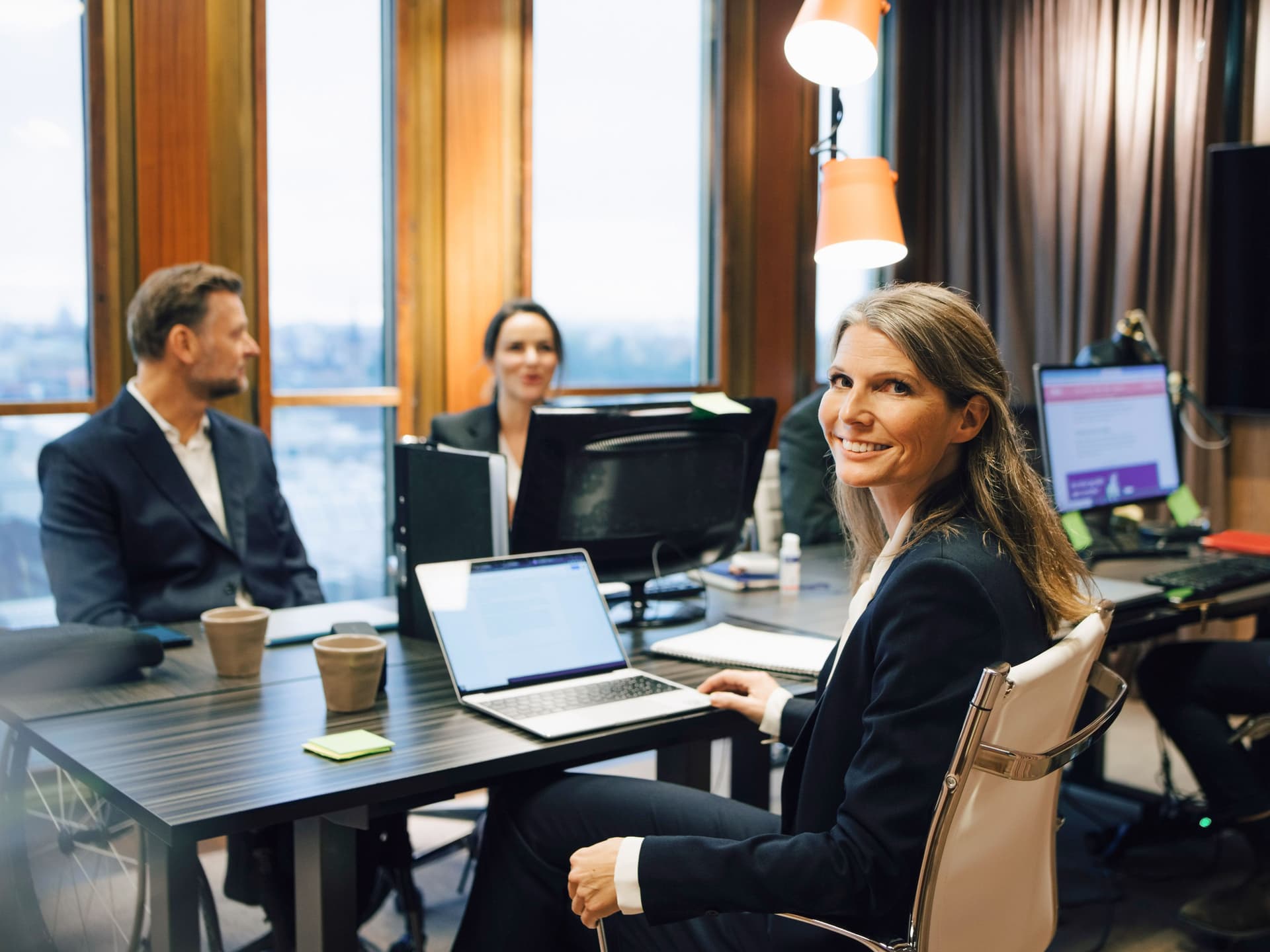 Eine Frau in Business-Kleidung dreht sich während eines Meetings mit drei Männern und einer Frau in einem Konferenzraum lächelnd in die Kamera. Sie hat einen aufgeklappten Laptop vor sich.
