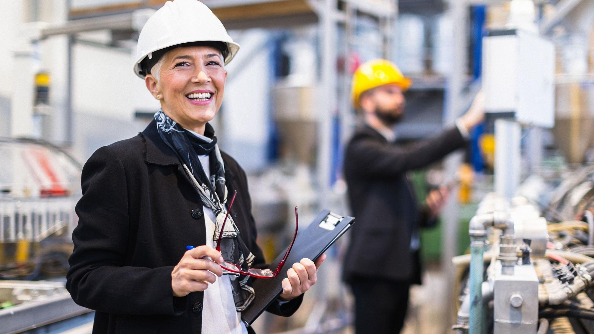 Eine Frau steht mit einem Helm in einer Fabrik und lächelt in die Kamera. Sie hat eine Brille und einen Ordner in den Händen.
