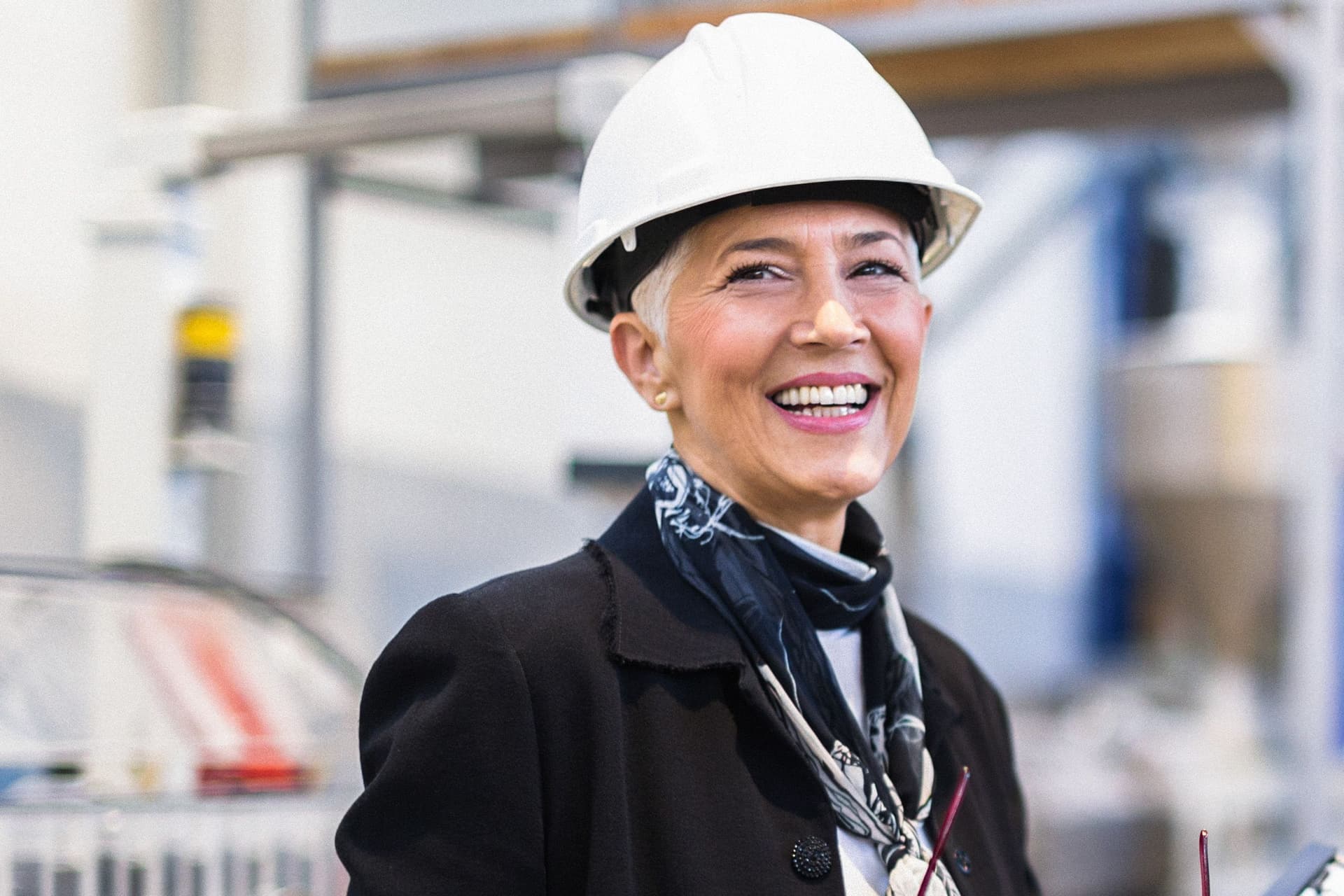 Eine Frau steht mit einem Helm in einer Fabrik und lächelt in die Kamera. Sie hat eine Brille und einen Ordner in den Händen.