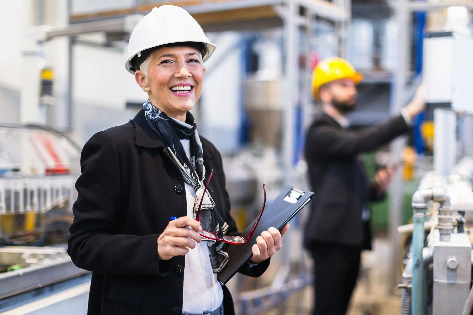 Eine Frau steht mit einem Helm in einer Fabrik und lächelt in die Kamera. Sie hat eine Brille und einen Ordner in den Händen.