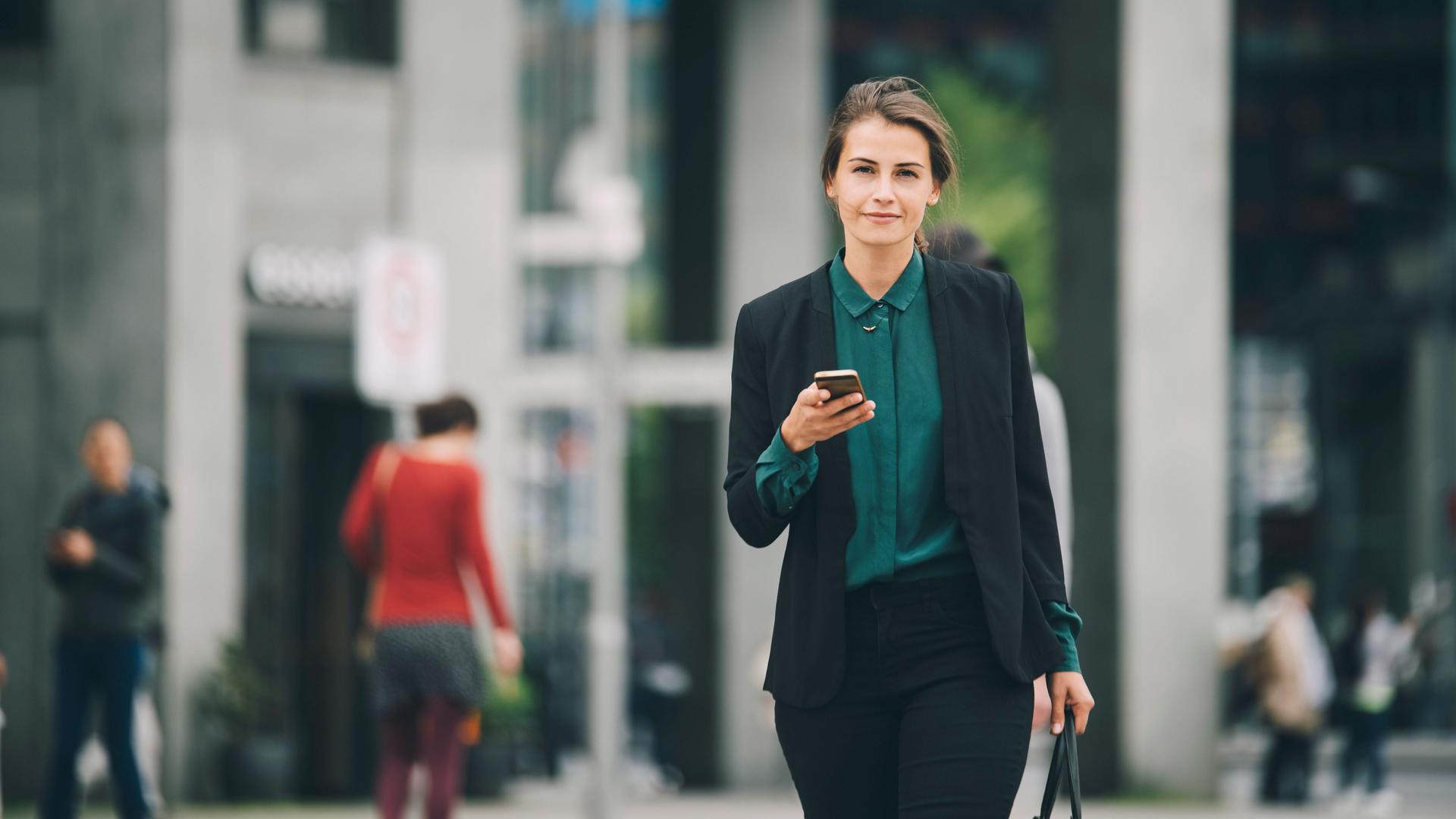 Junge Frau in Business-Kleidung mit Mobiltelefon in einer Hand und einer Tasche in der anderen, läuft auf einer Straße und blickt in die Kamera.