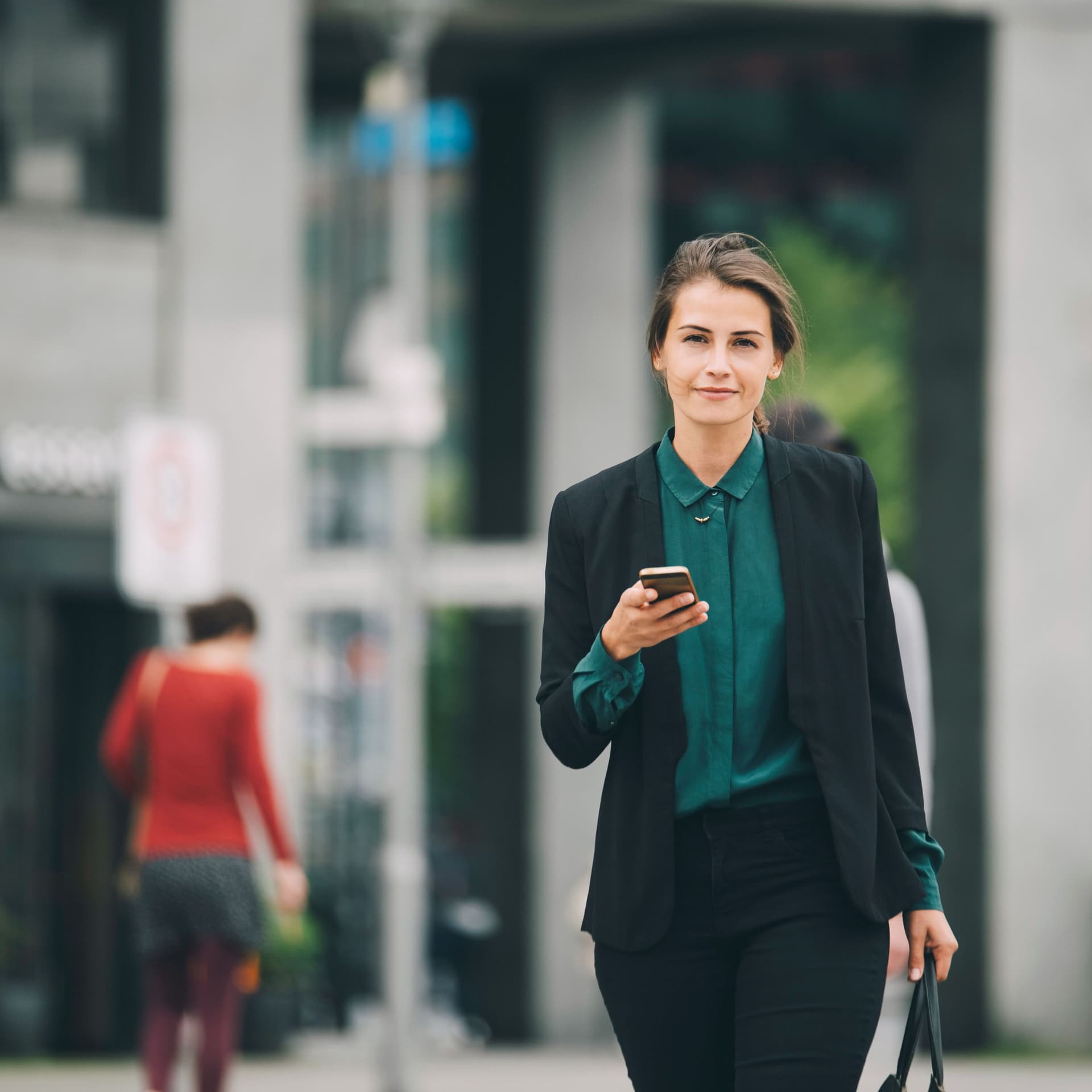 Junge Frau in Business-Kleidung mit Mobiltelefon in einer Hand und einer Tasche in der anderen, läuft auf einer Straße und blickt in die Kamera.