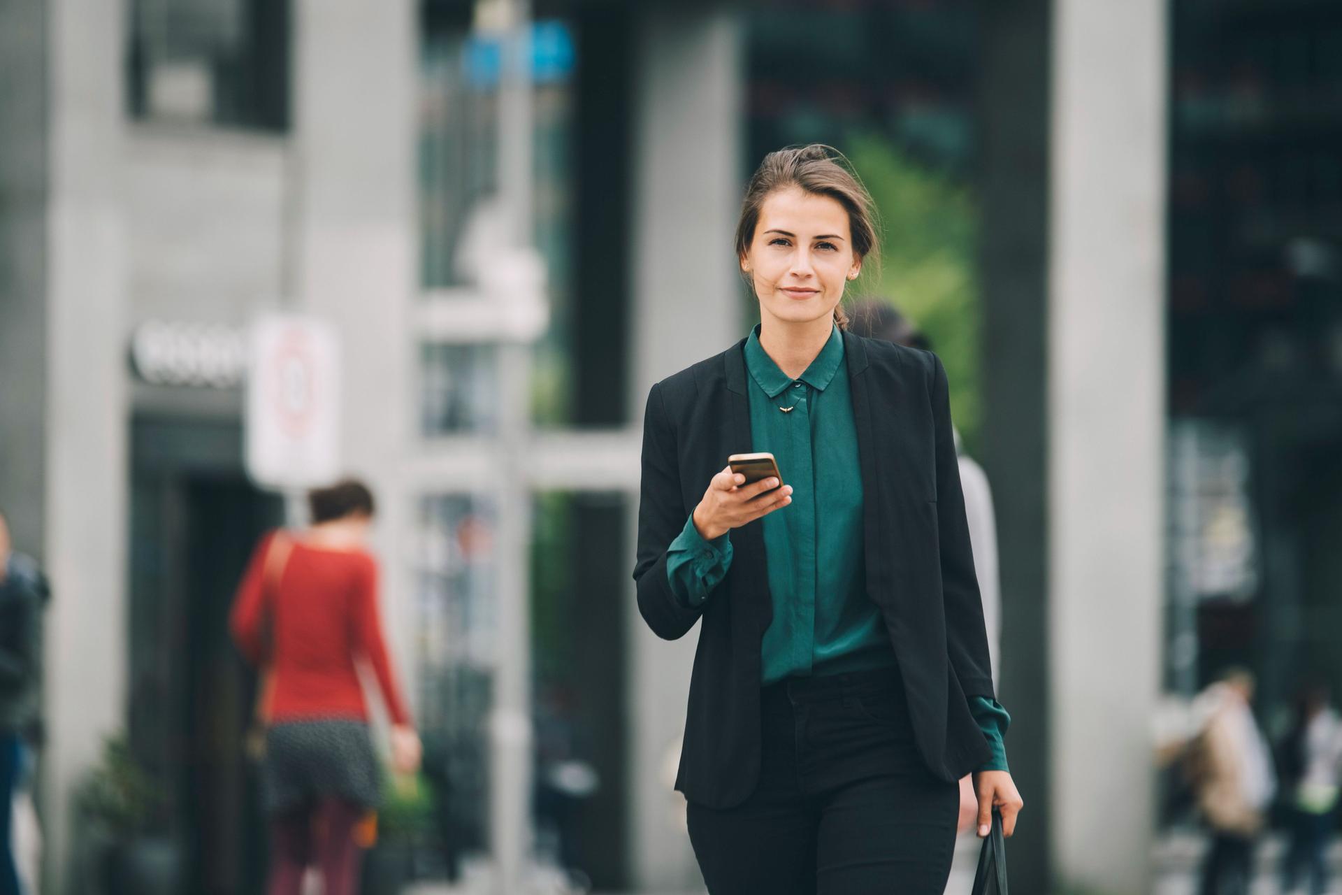 Junge Frau in Business-Kleidung mit Mobiltelefon in einer Hand und einer Tasche in der anderen, läuft auf einer Straße und blickt in die Kamera.