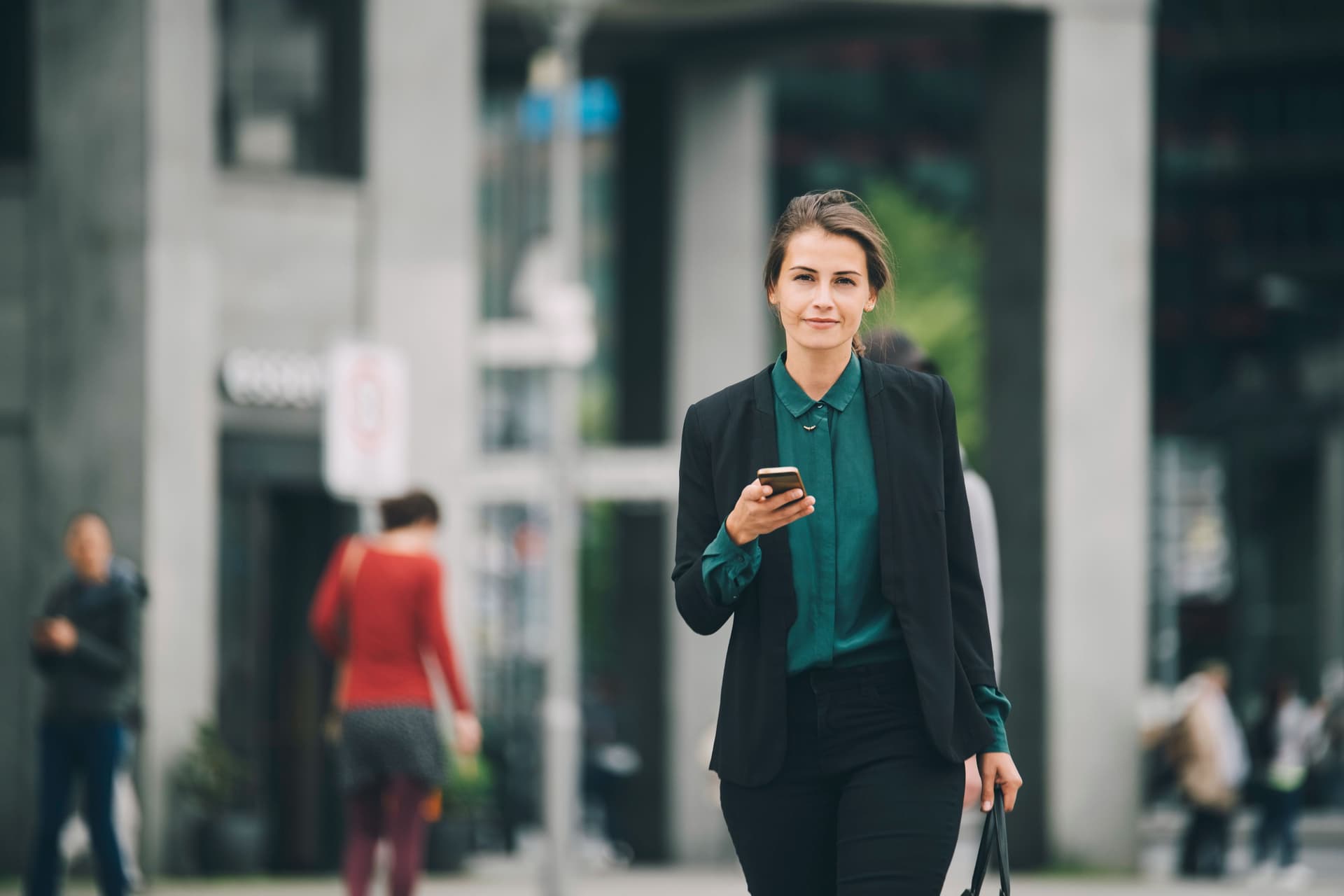 Junge Frau in Business-Kleidung mit Mobiltelefon in einer Hand und einer Tasche in der anderen, läuft auf einer Straße und blickt in die Kamera.