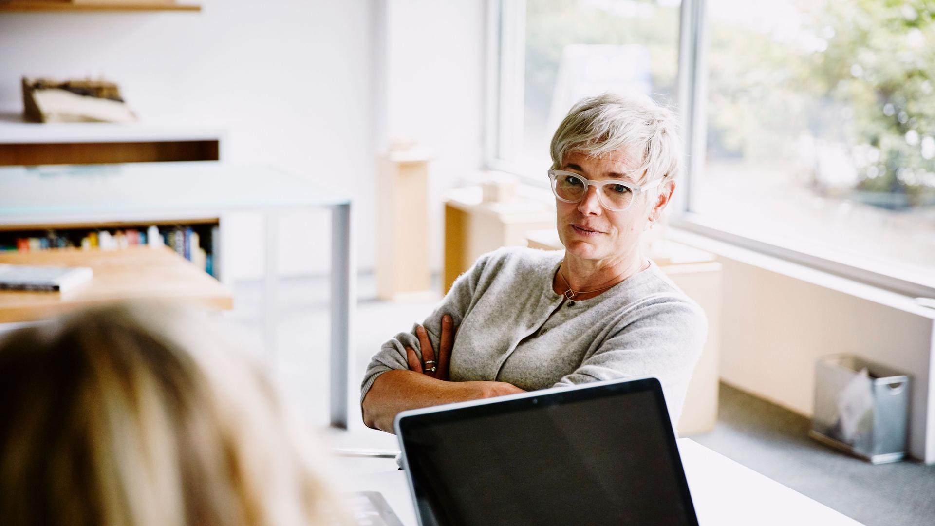 Eine ältere Frau mit Brille sitzt mit verschränkten Armen an einem Konferenztisch in einem Büro mit einer anderen Frau, dessen Hinterkopf man sieht.