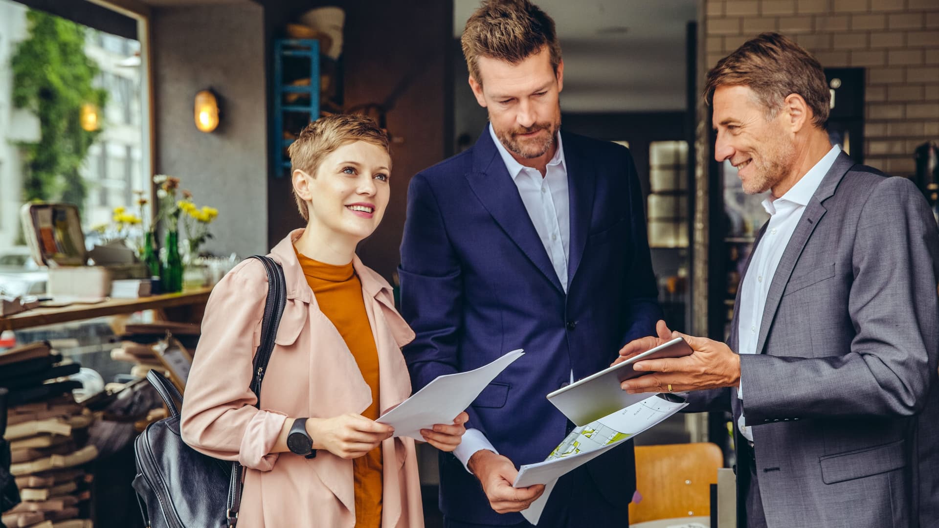 Geschäftsmann zeigt einer Frau und einem Mannn, die Grundrisszeichnungen in der Hand halten, etwas auf einem Tablet-Computer.