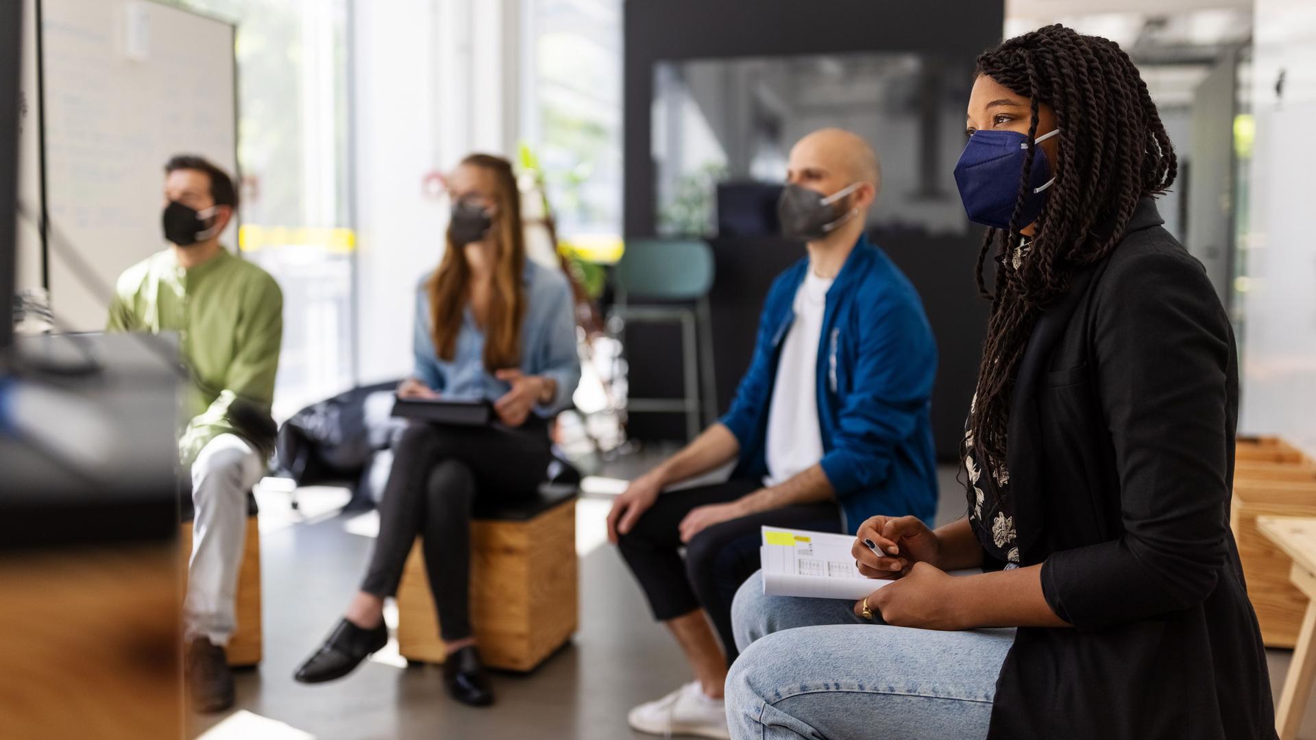 Frauen und Männer sitzen mit Gesichtsmasken auf Holzhockern in einem Büro. Sie gucken alle in eine Richtung auf einen Bildschirm.