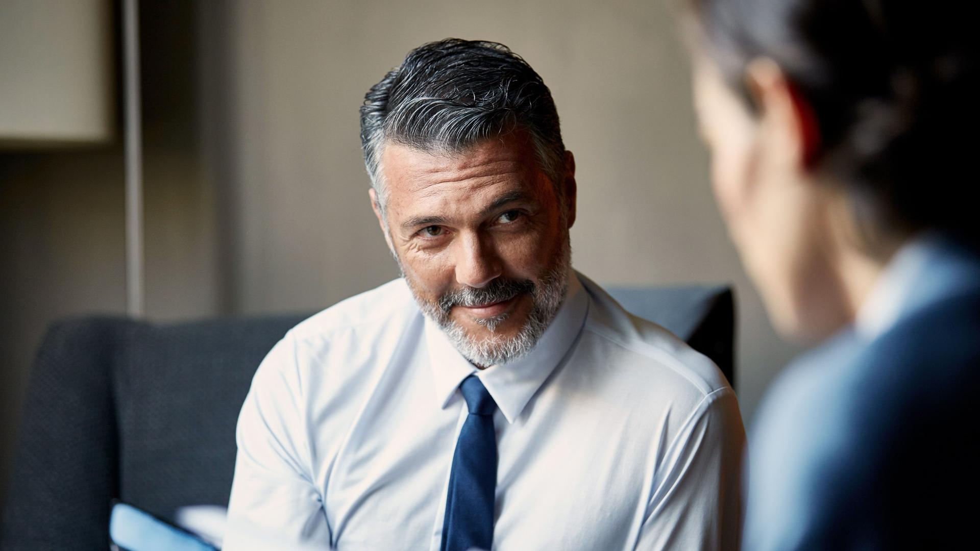 Close-Up eines Mannes in Hemd und Krawatte, der auf einem Sessel sitzt und seine Gesprächspartnerin lächelnd anblickt.