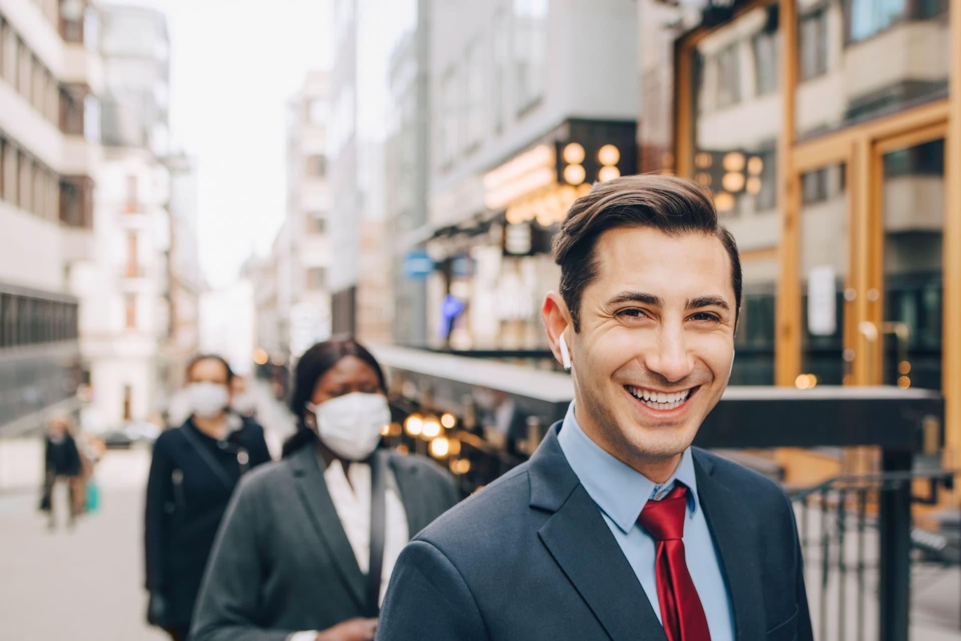 Ein junger Mann in Business-Kleidung läuft auf einer urbanen Straße mit kabellosem Kopfhörer im Ohr und blickt dabei lächelnd in die Kamera,
