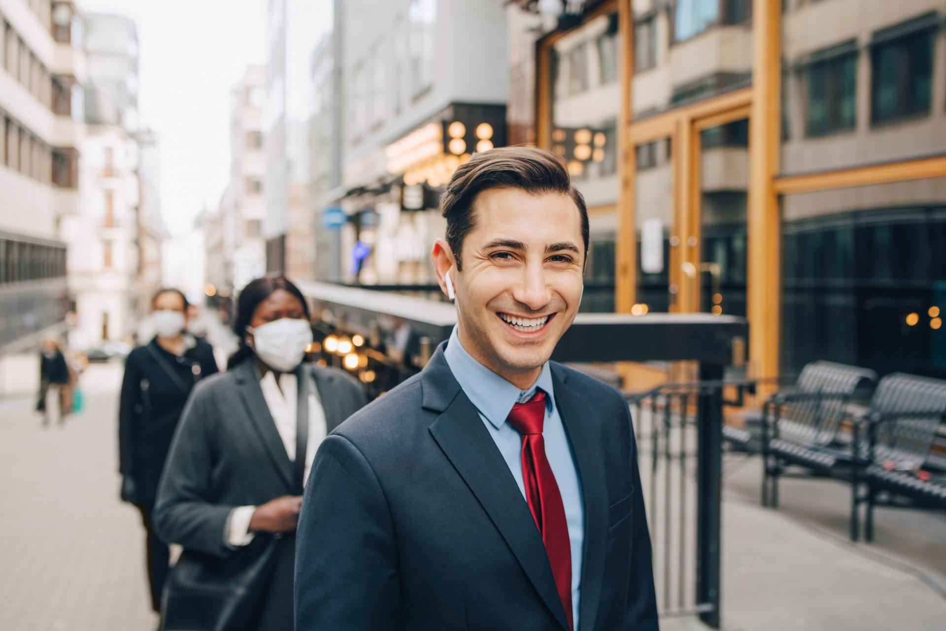 Ein junger Mann in Business-Kleidung läuft auf einer urbanen Straße mit kabellosem Kopfhörer im Ohr und blickt dabei lächelnd in die Kamera, 