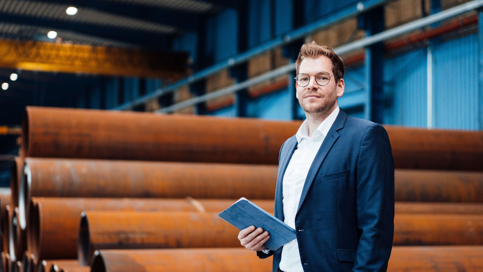 Ein Mann im Anzug steht mit einem Tablet in einer Fabrikhalle vor grossen Stahlrohren.