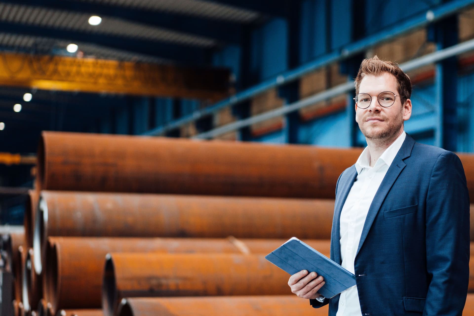 Ein Mann im Anzug steht mit einem Tablet in einer Fabrikhalle vor grossen Stahlrohren.