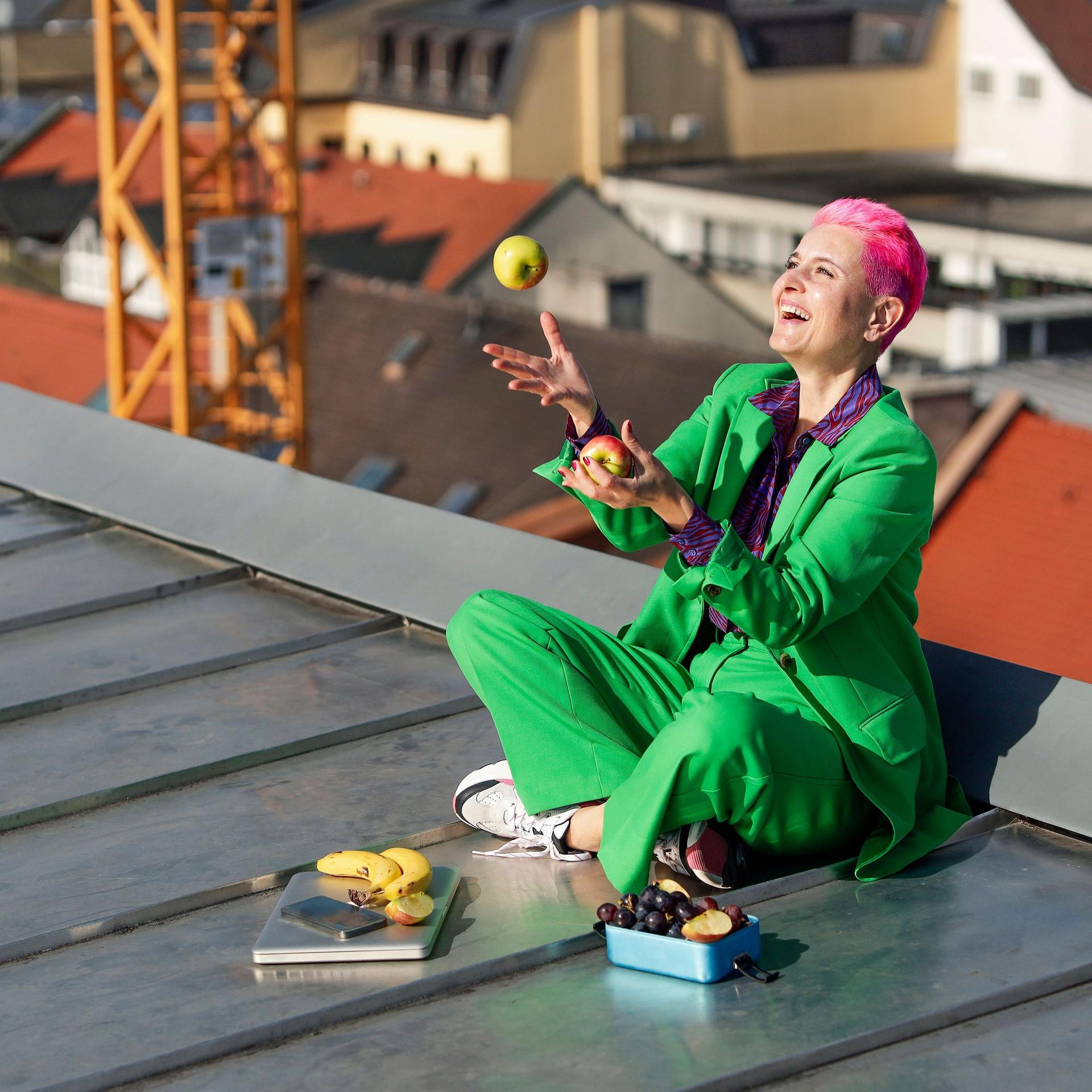 Lachende Frau sitzt auf einem Dach und jongliert mit zwei Äpfeln. Vor ihr liegt ein zugeklappter Laptop und mehr Obst in einer Dose.