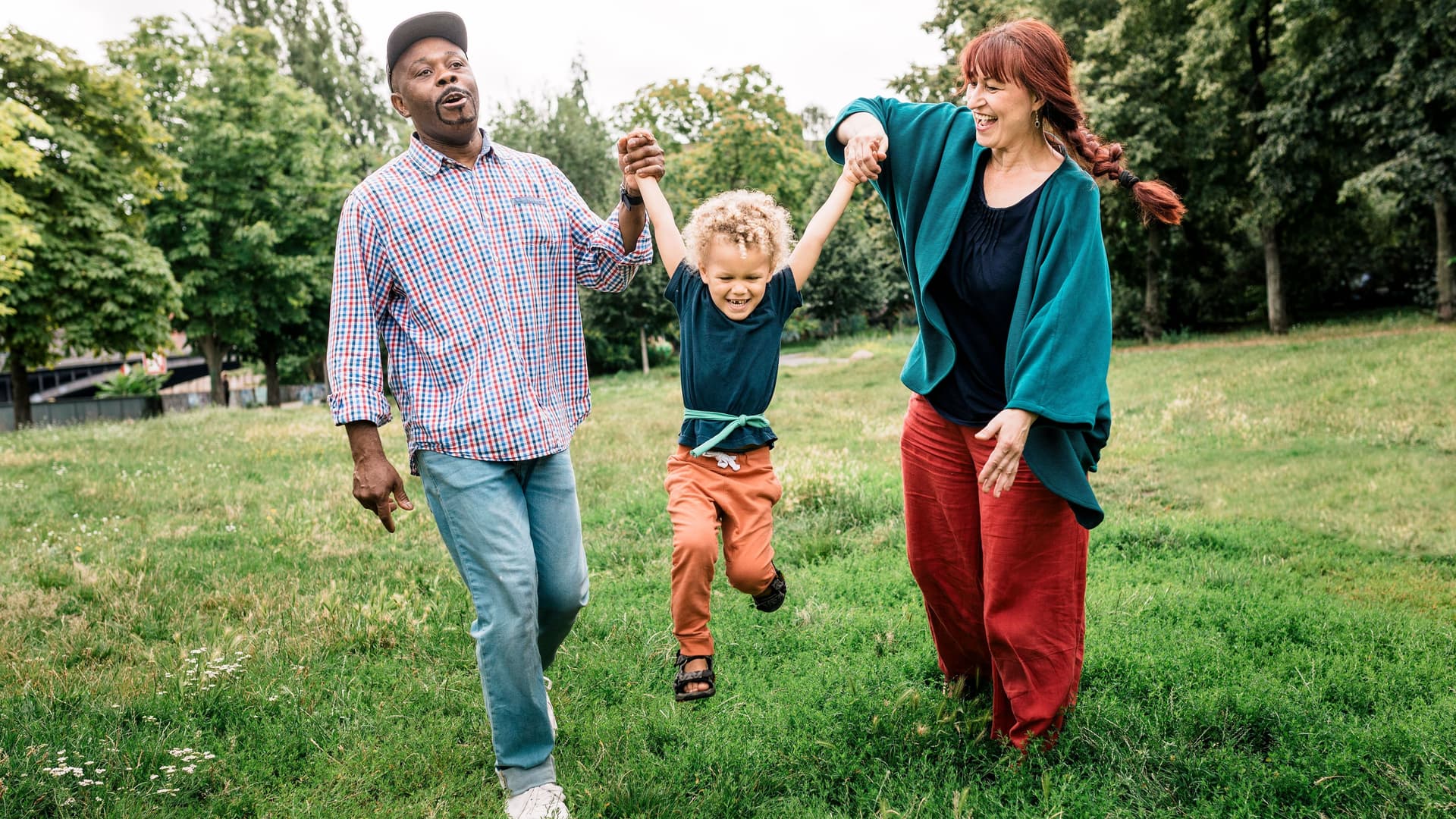 Ein mittelalter Mann und eine mittelalte Frau schwingen einen kleinen Jungen an den Händen in einem Park. Alle drei lächeln.