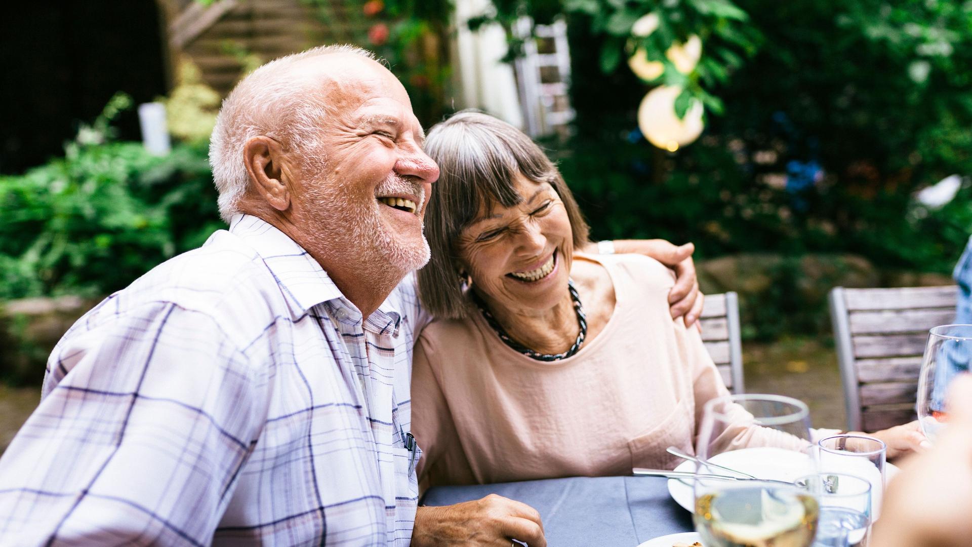 Ein älteres Paar, das sich umarmt und mit der Familie in einem Innenhof eine Mahlzeit im Freien genießt.
