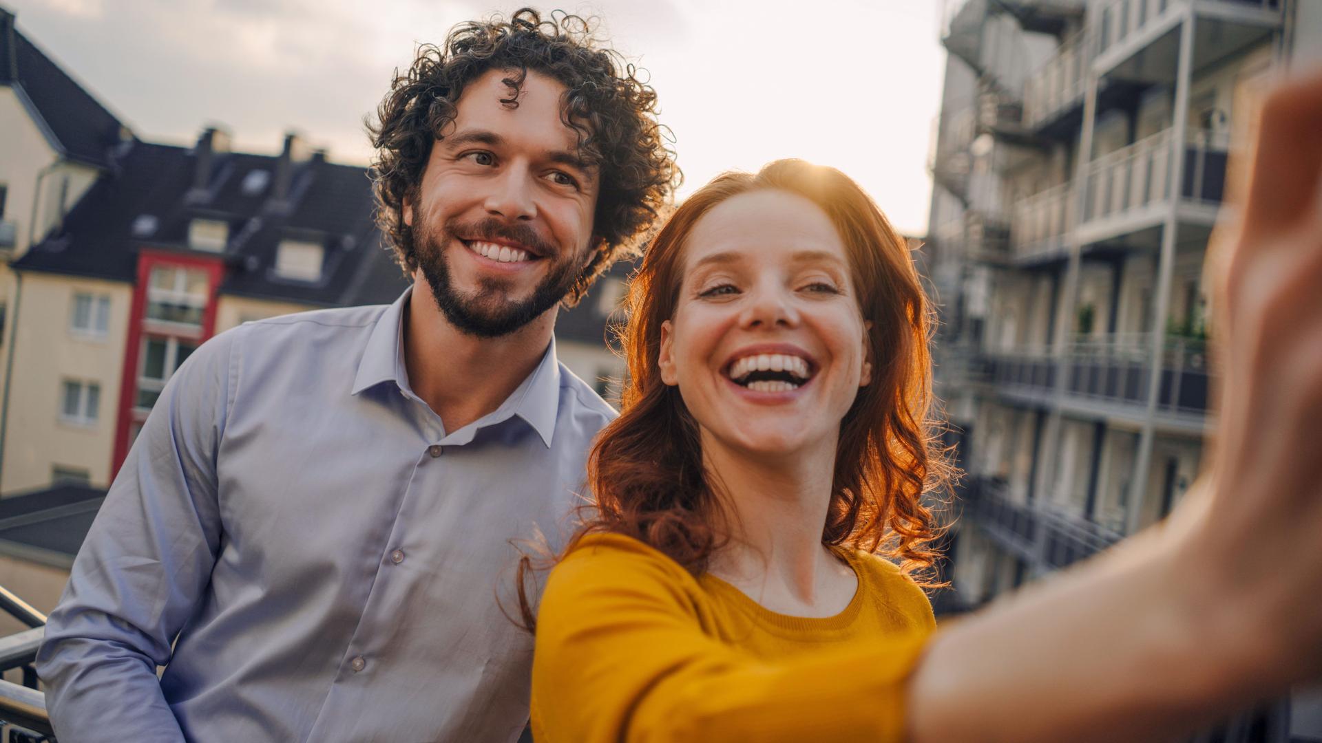 Ein Mann und eine Frau machen auf einem Balkon ein Selfie. Sie lachen ausgelassen in die Kamera. Im Hintergrund sind weitere Häuser zu sehen.