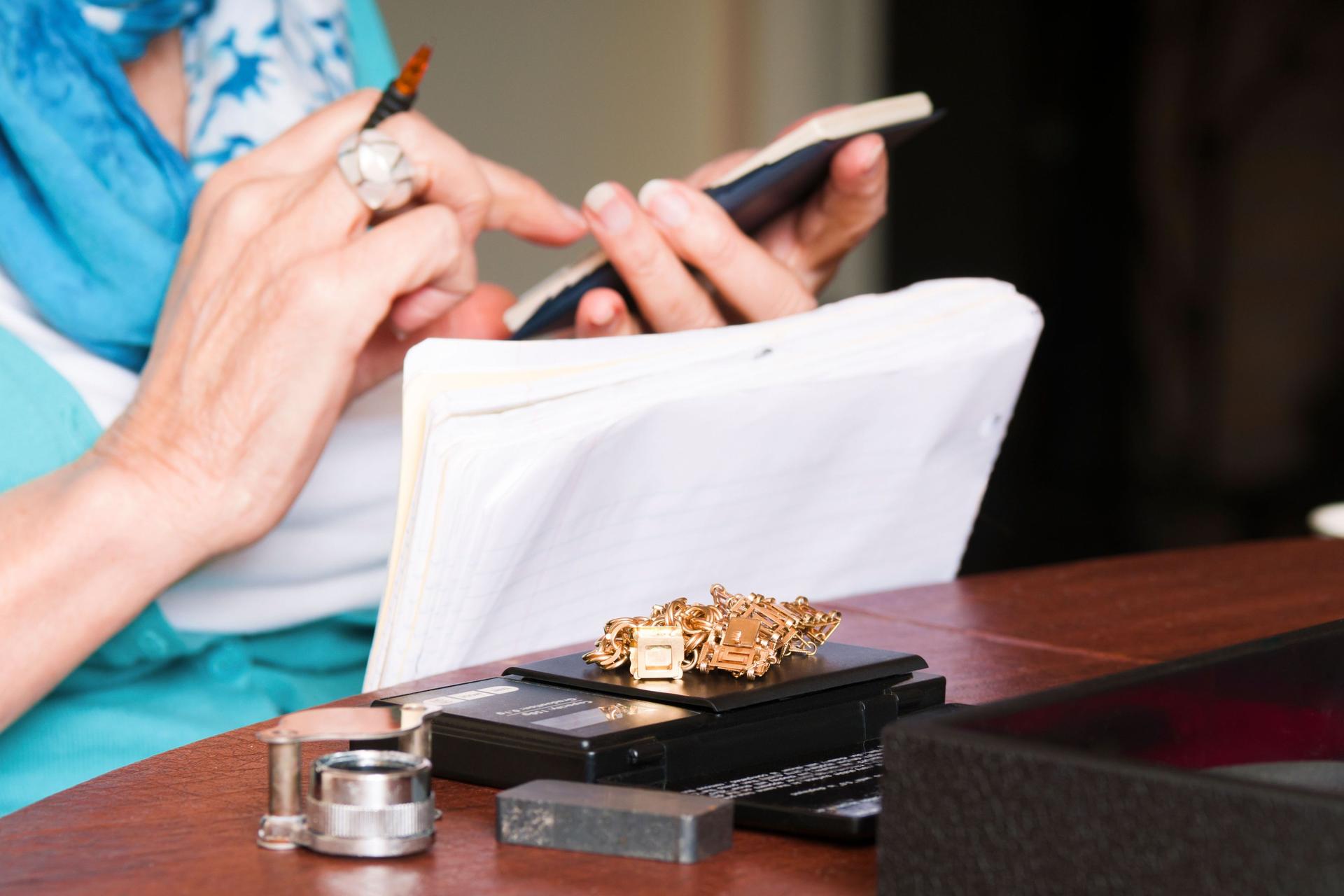 Nahaufnahme von Frauenhänden mit einem Taschenrechner und Stift. Vor ihr liegt auf einem Holztisch eine Schmuckwaage mit Goldstücken. 