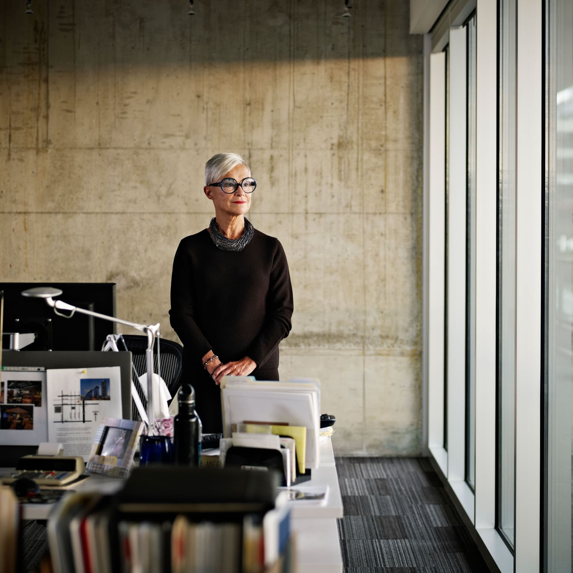Grauhaarige Frau mit schwarzer Brille schaut aus dem Fenster in einem moderenen lichtdurchfluteten Grossraumbuero.