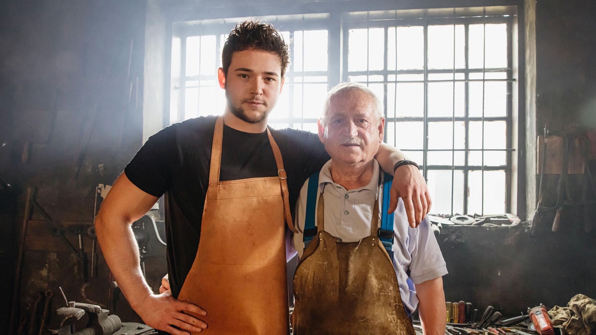 Porträt von einem jungen und einem alten Mann mit Schürzen, die Arm in Arm in einer Schmiede stehen.