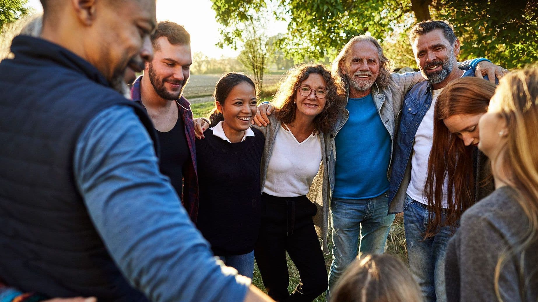 Eine Gruppe von Menschen verschieden Alters stehen in den Abendstunden im Grünen und umarmen sich im Kreis.