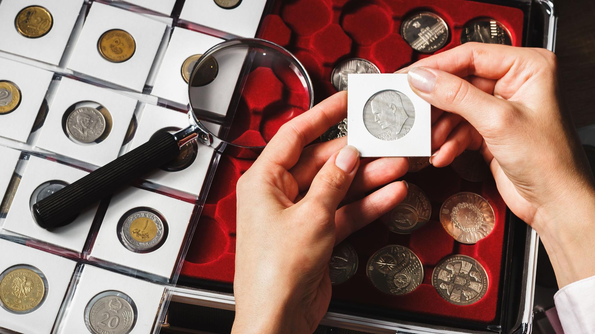 Nahaufnahme zweier Hände die eine eingepackte Sammelmünze halten. Links eine Lupe und Seite mit Münzen in einzelnen kleinen Taschen. Rechts eine Schachtel mit Sammelmünzen. 