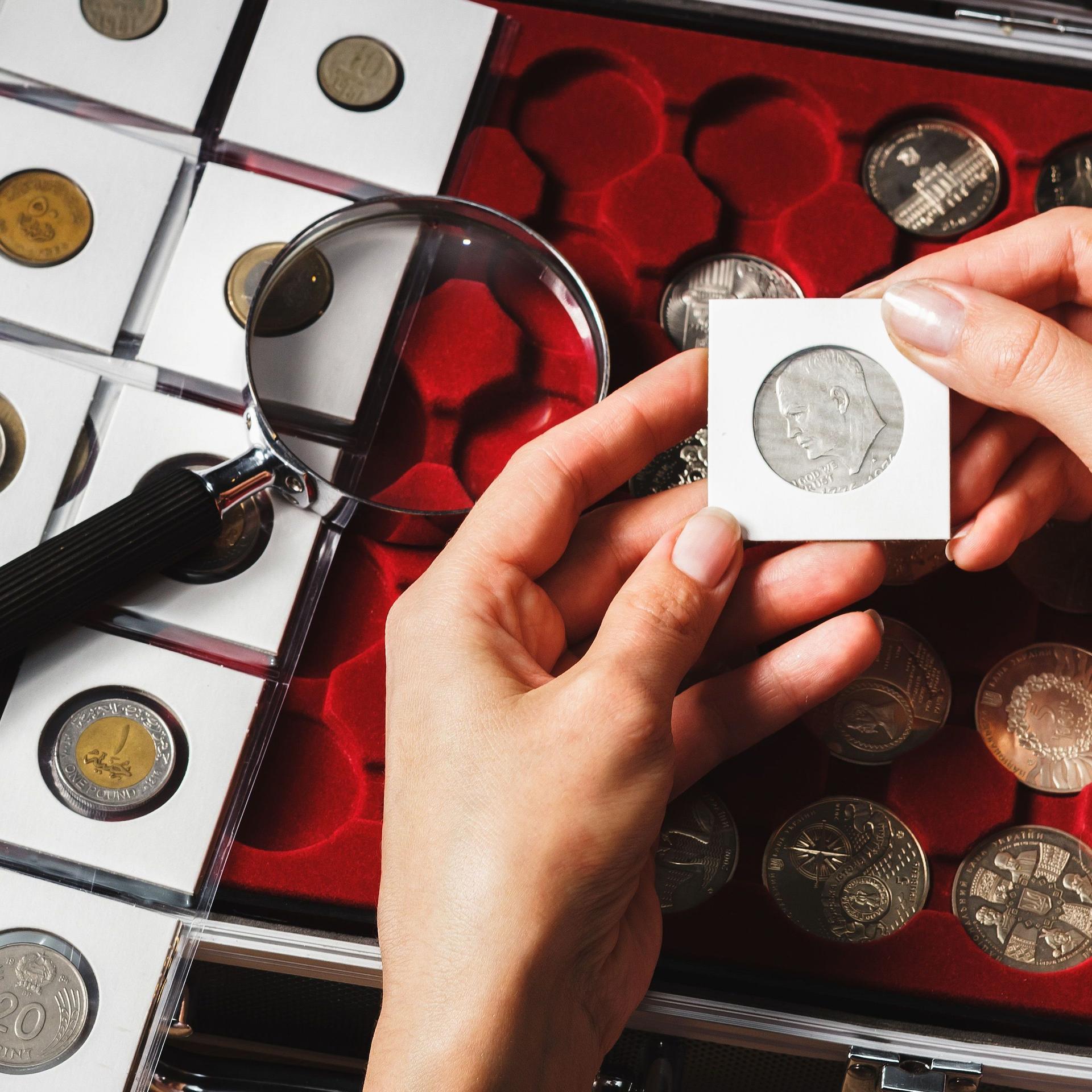Nahaufnahme zweier Hände die eine eingepackte Sammelmünze halten. Links eine Lupe und Seite mit Münzen in einzelnen kleinen Taschen. Rechts eine Schachtel mit Sammelmünzen. 