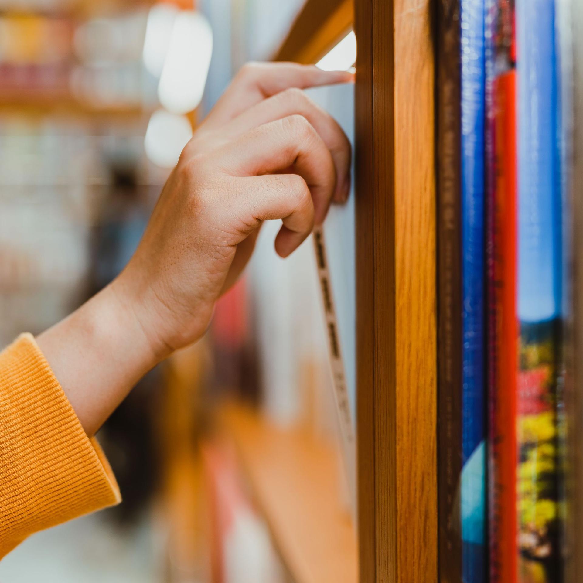 Seitliche Nahaufnahme von einem Arm mit orange farbenen Pullover. Die Hand zieht ein Buch aus einem Bücherregal.