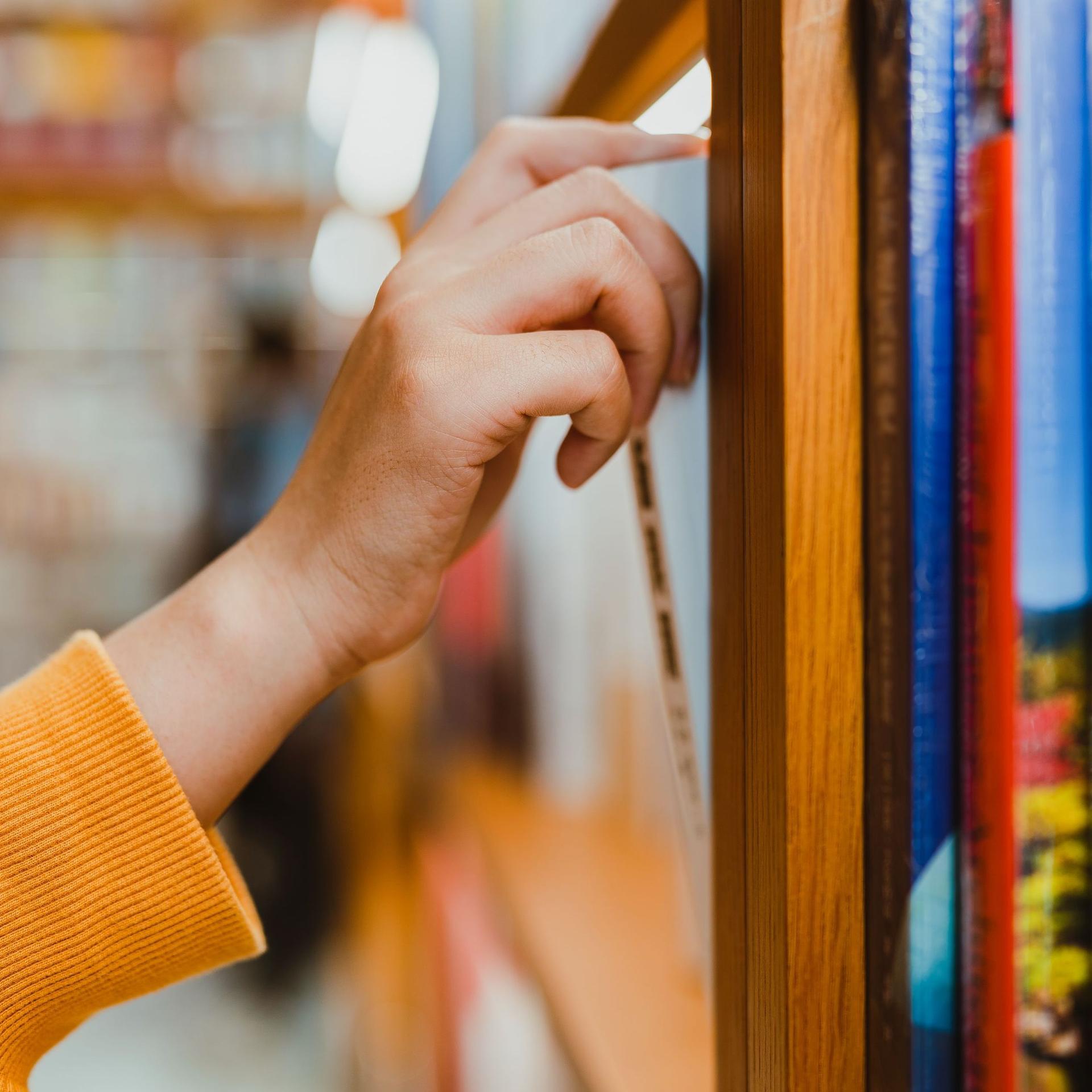 Seitliche Nahaufnahme von einem Arm mit orangefarbenen Pullover. Die Hand zieht ein Buch aus einem Bücherregal.