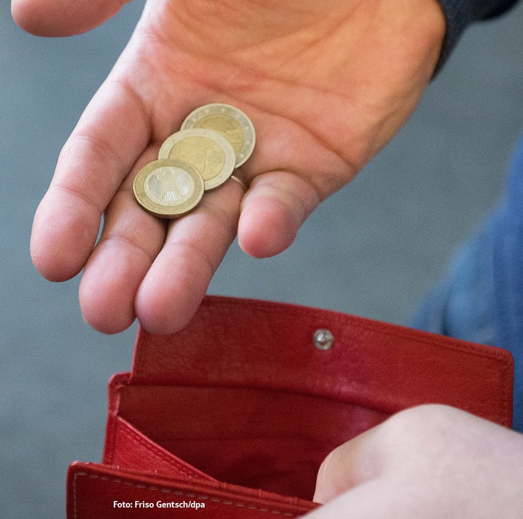 Eine Hand hält wenige Euromünzen über einer offenen roten Geldbörse, was Armut oder knappe Finanzen symbolisiert.