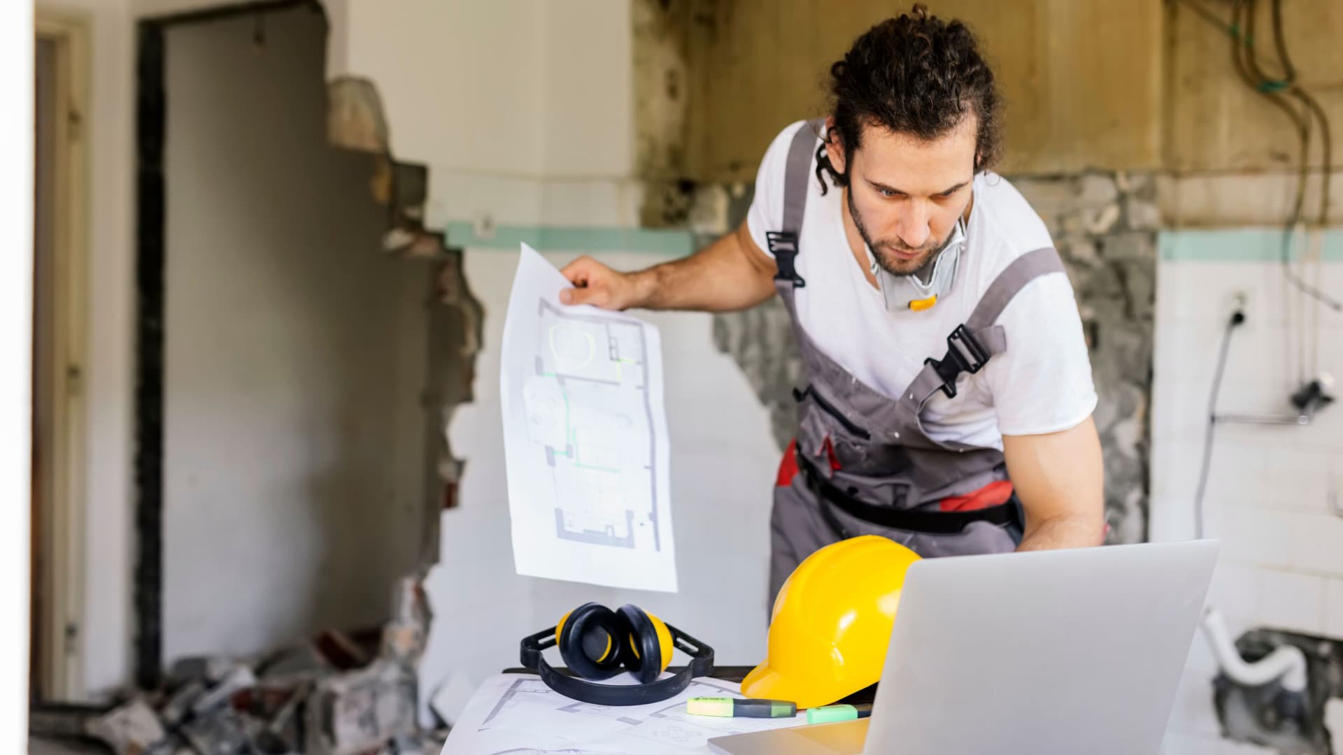 Arbeiter, der sich ein Architekturprojekt auf Papier und Laptop ansieht. Hinter ihm ist eine eingerissene Wand.