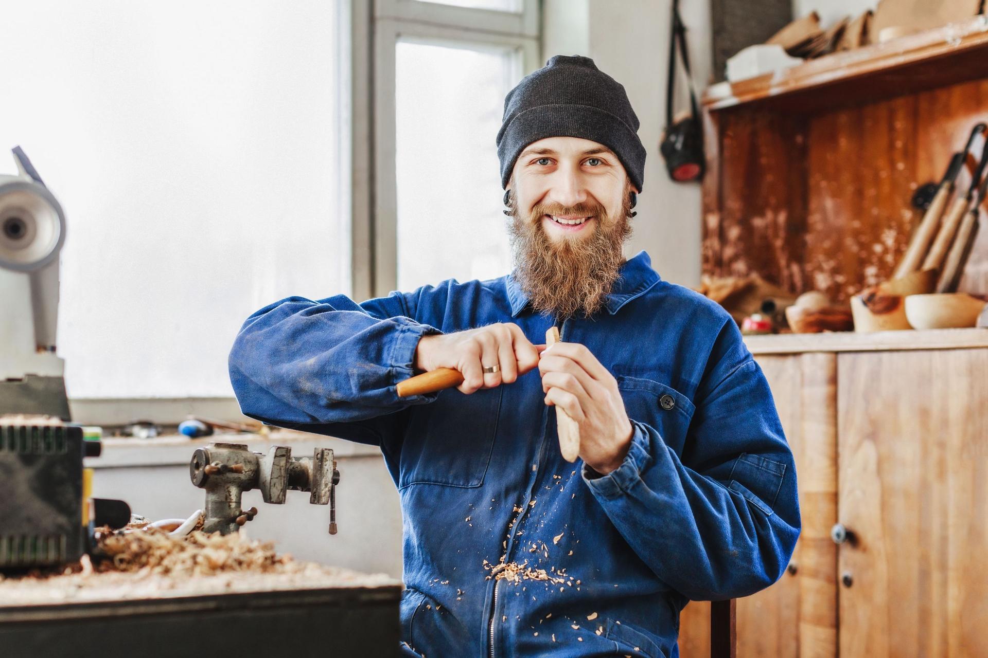 Lächelnder Mann mit Bart in blauem Jeansanzug und schwarzem Hut, der Holzlöffel mit Instrumenten schnitzt und in die Kamera schaut.