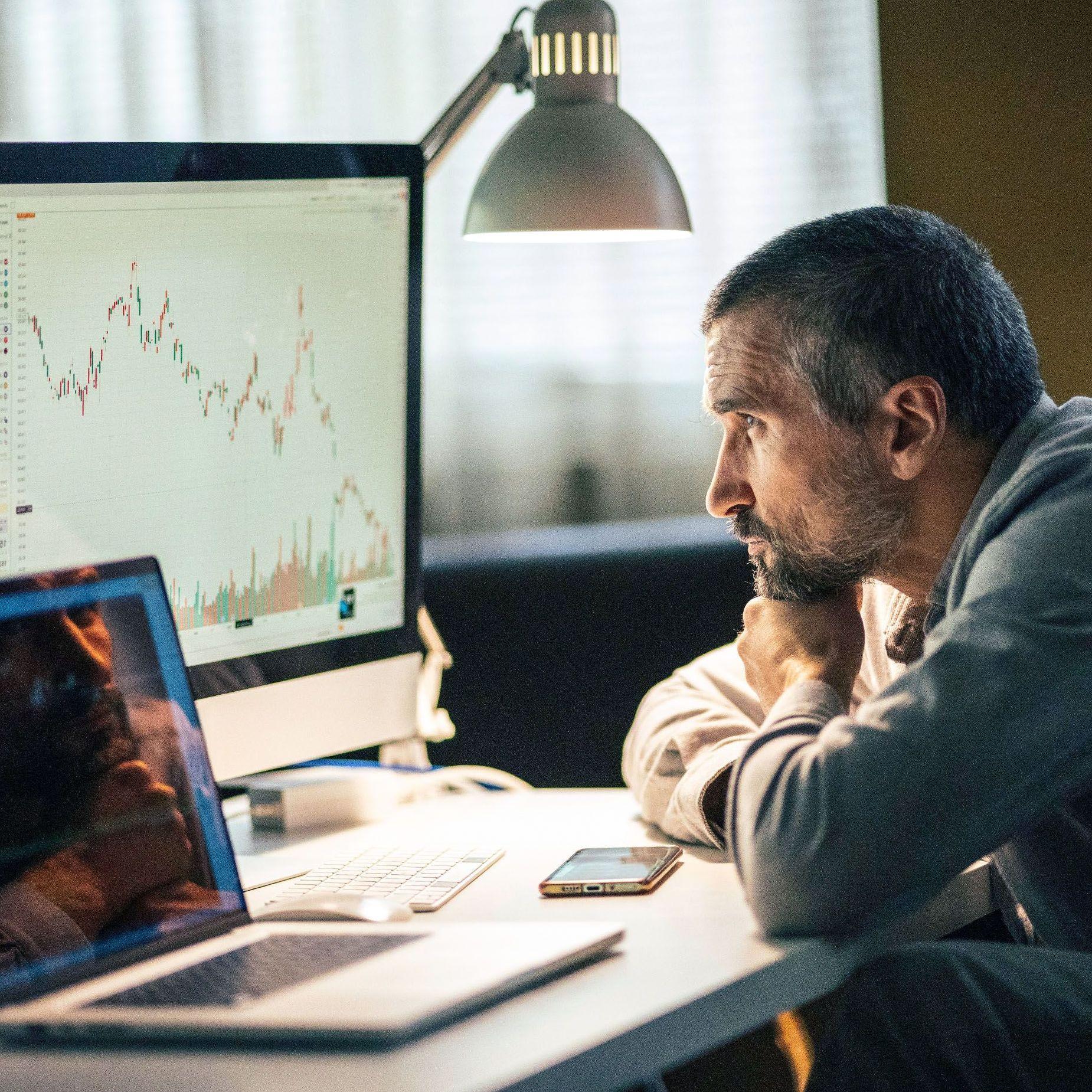 Ein Mann mittleren Alters schaut ernsthaft, konzentriert, sein Kinn auf seine Hände gestützt, auf die Aktienkurven mehrerer Bildschirme auf dem Schreibtisch vor ihm. 