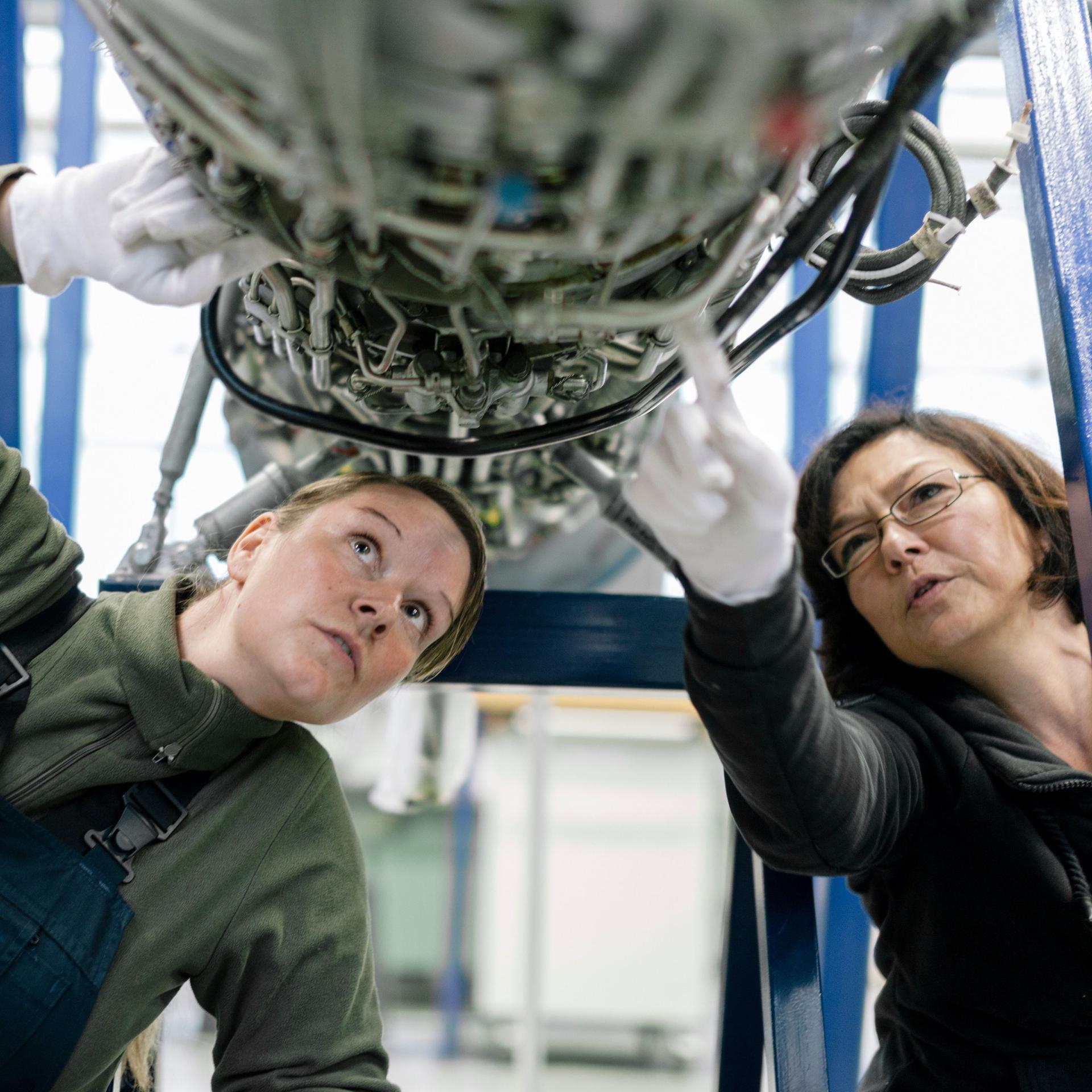 Zwei Mechanikerinnen untersuchen ein Flugzeugtriebwerk. Sie schauen von unten auf eine Vielzahl von Kabeln.