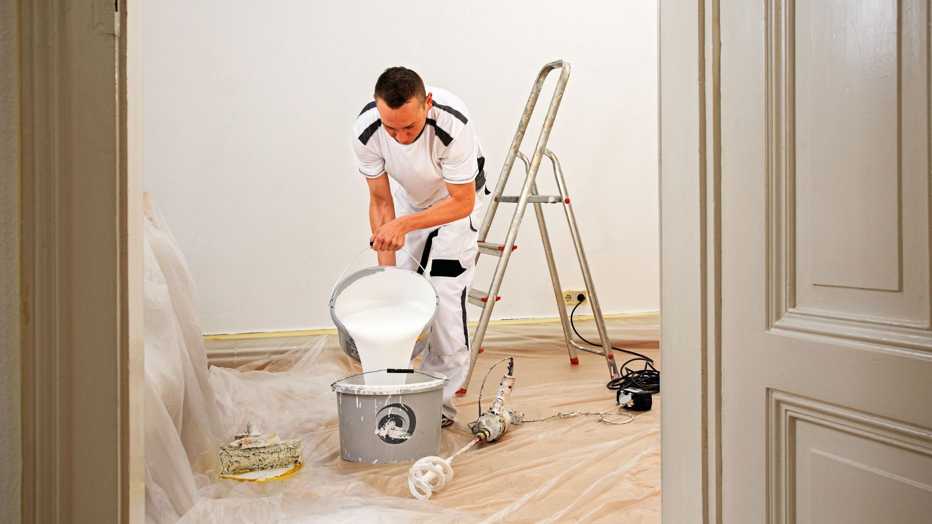 Maler steht in einem Raum mit einer Leiter und kippt Farbe in einen Topf.