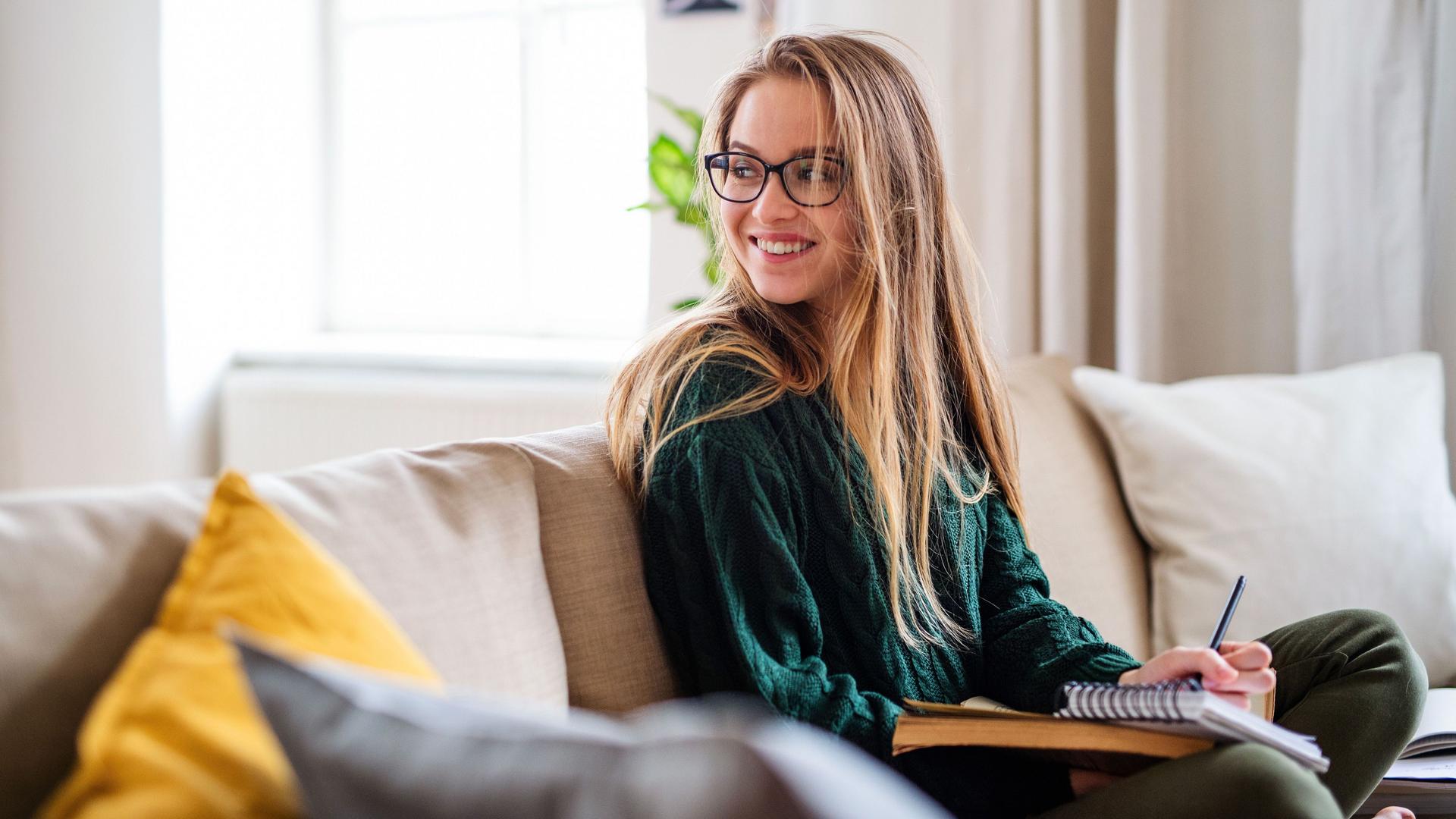 Eine lachende junge Frau mit Brille, die auf dem Sofa sitzt und studiert. Sie guckt über ihre Schulter nach hinten.