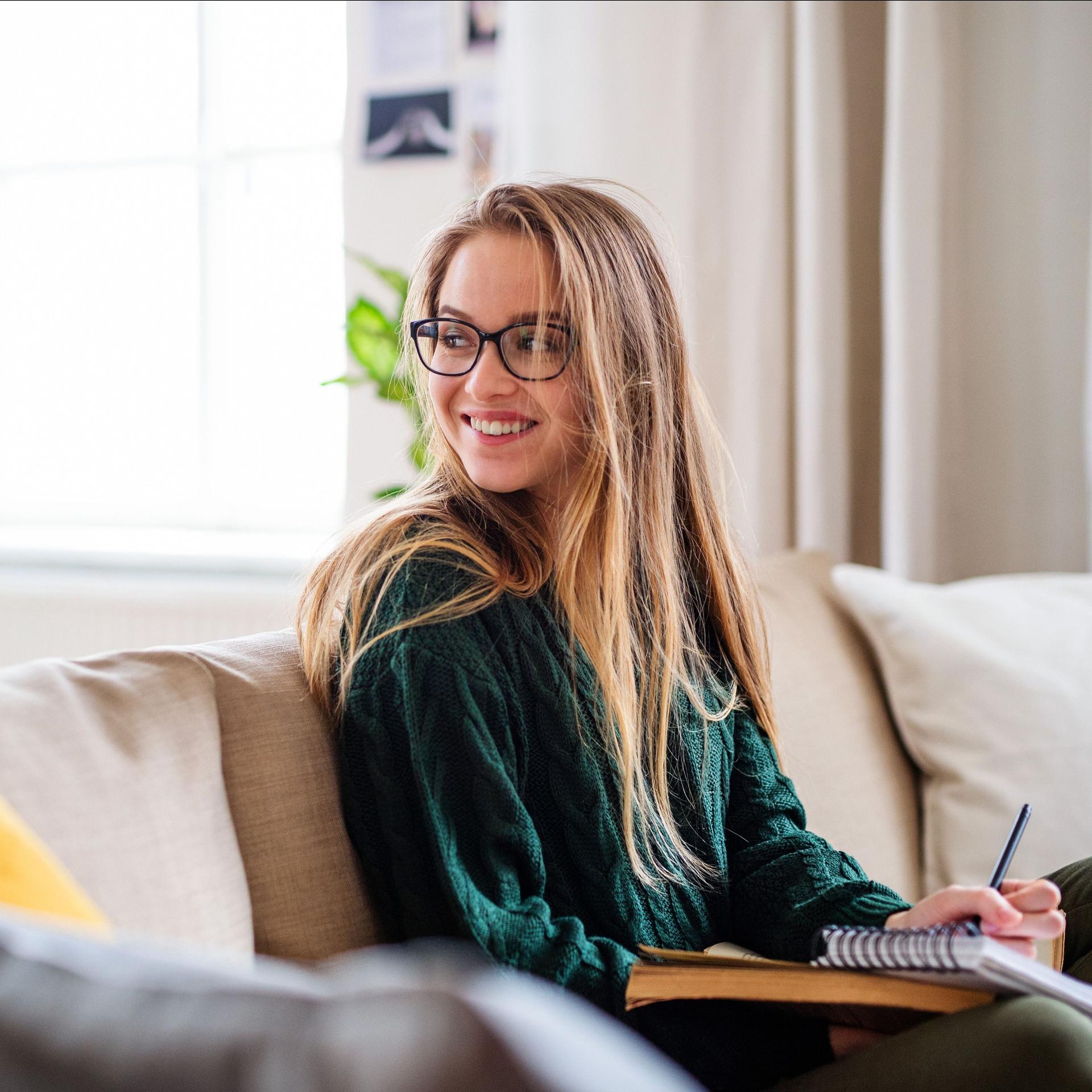 Eine lachende junge Frau mit Brille, die auf dem Sofa sitzt und studiert. Sie guckt über ihre Schulter nach hinten.