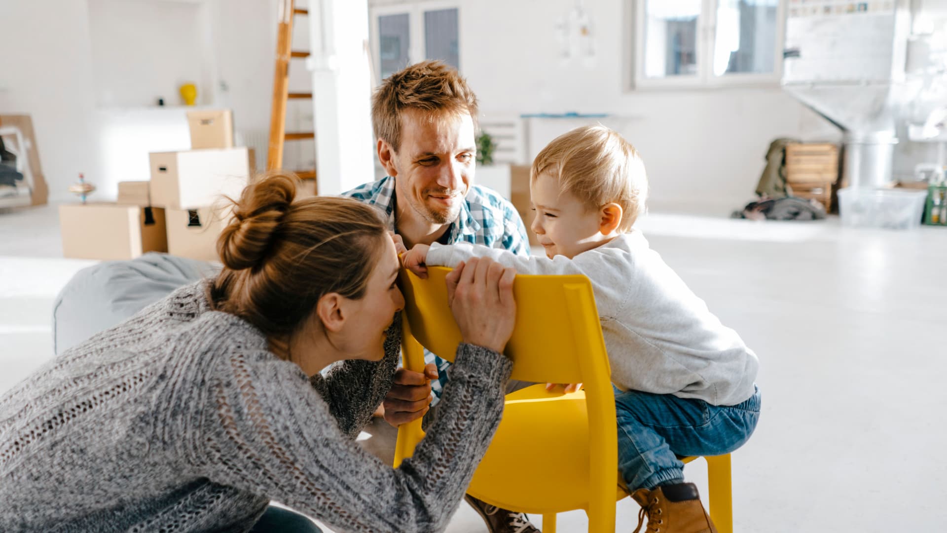 Junge Familie in einem modernen noch nicht fertig eingerichteten Raum. Die Mutter hockt vor einem gelben Stuhl und versteckt ihr Gesicht vor dem Kleinkind, was auf dem Stuhl sitzt.
