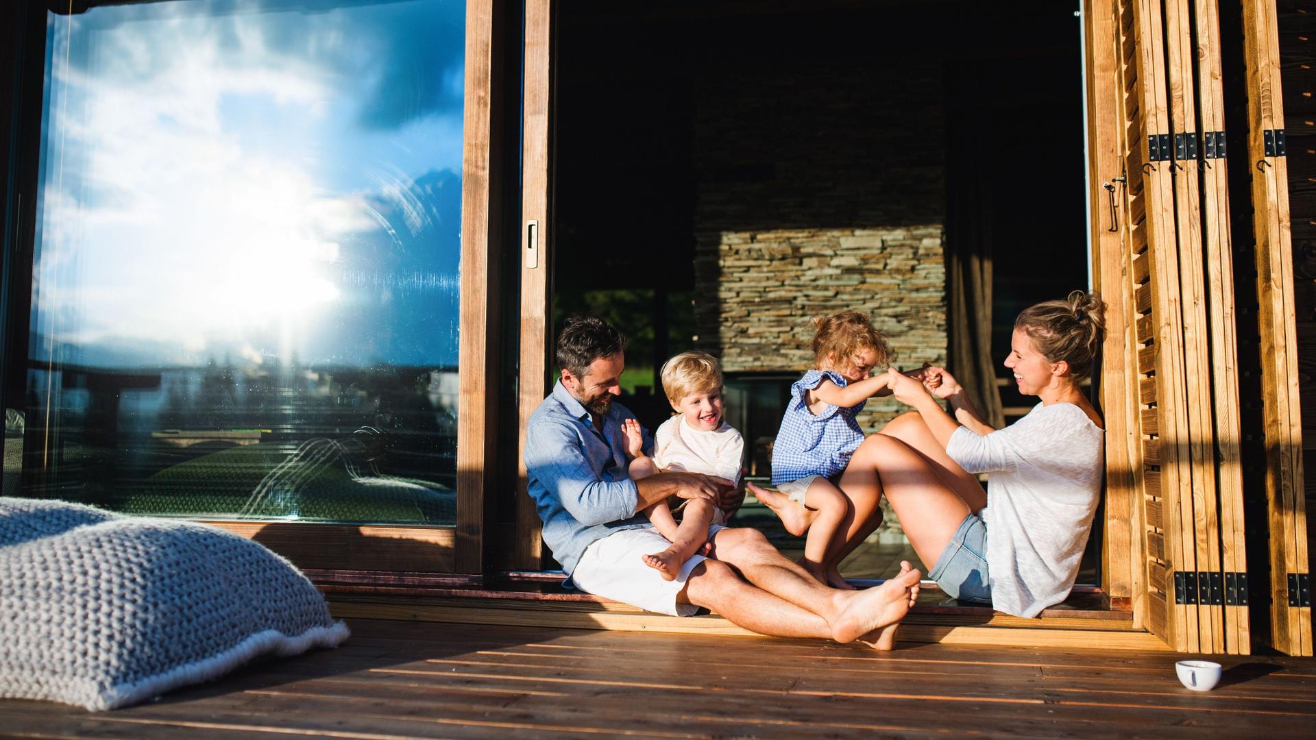 Familie mit kleinen Kindern, die auf dem Boden der Terrasse eines Holzhauses sitzen. Sie sind sommerlich gekleidet und die Sonne spiegelt sich in der Terassentür.