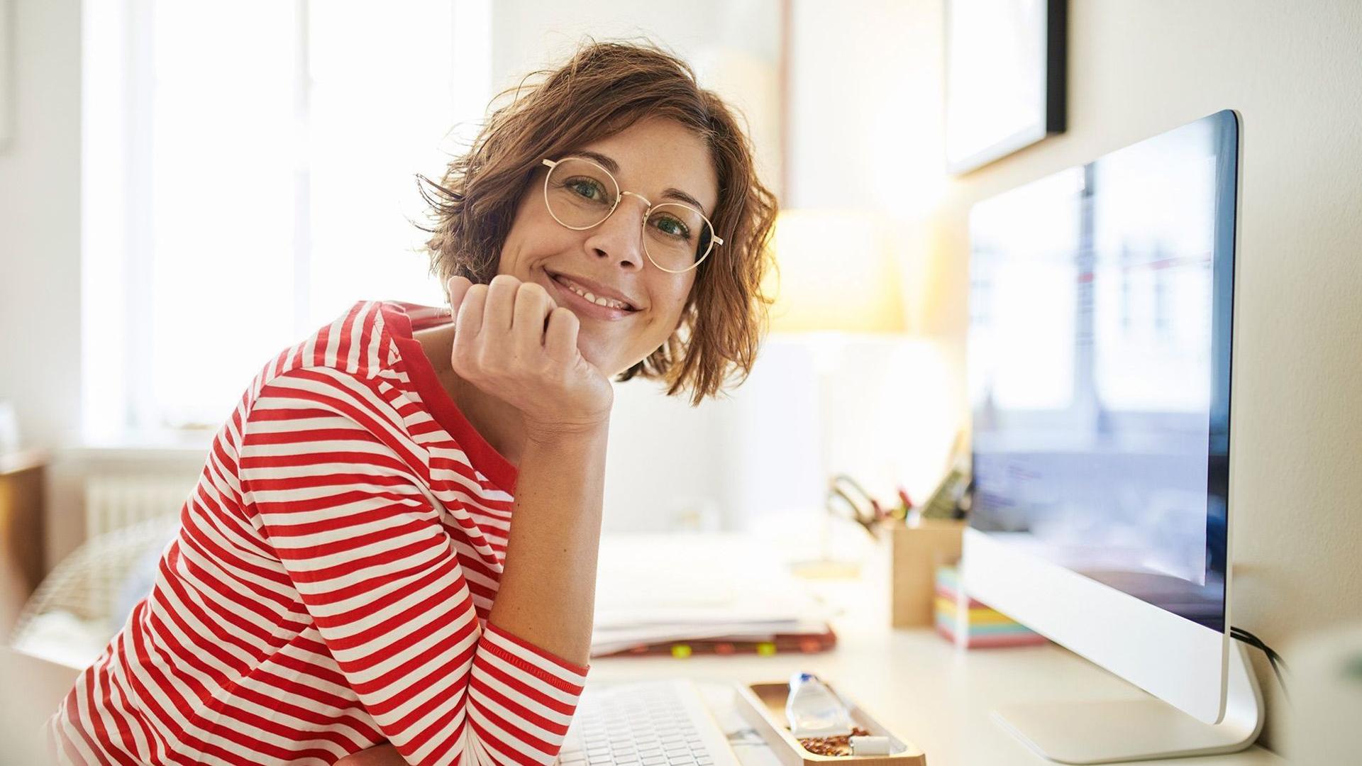 Eine junge Frau mit Brille und Ringelpulli sitzt an ihrem Schreibtisch. Sie hat ihren Kopf auf ihrer Hand abgestützt und lächelt in die Kamera