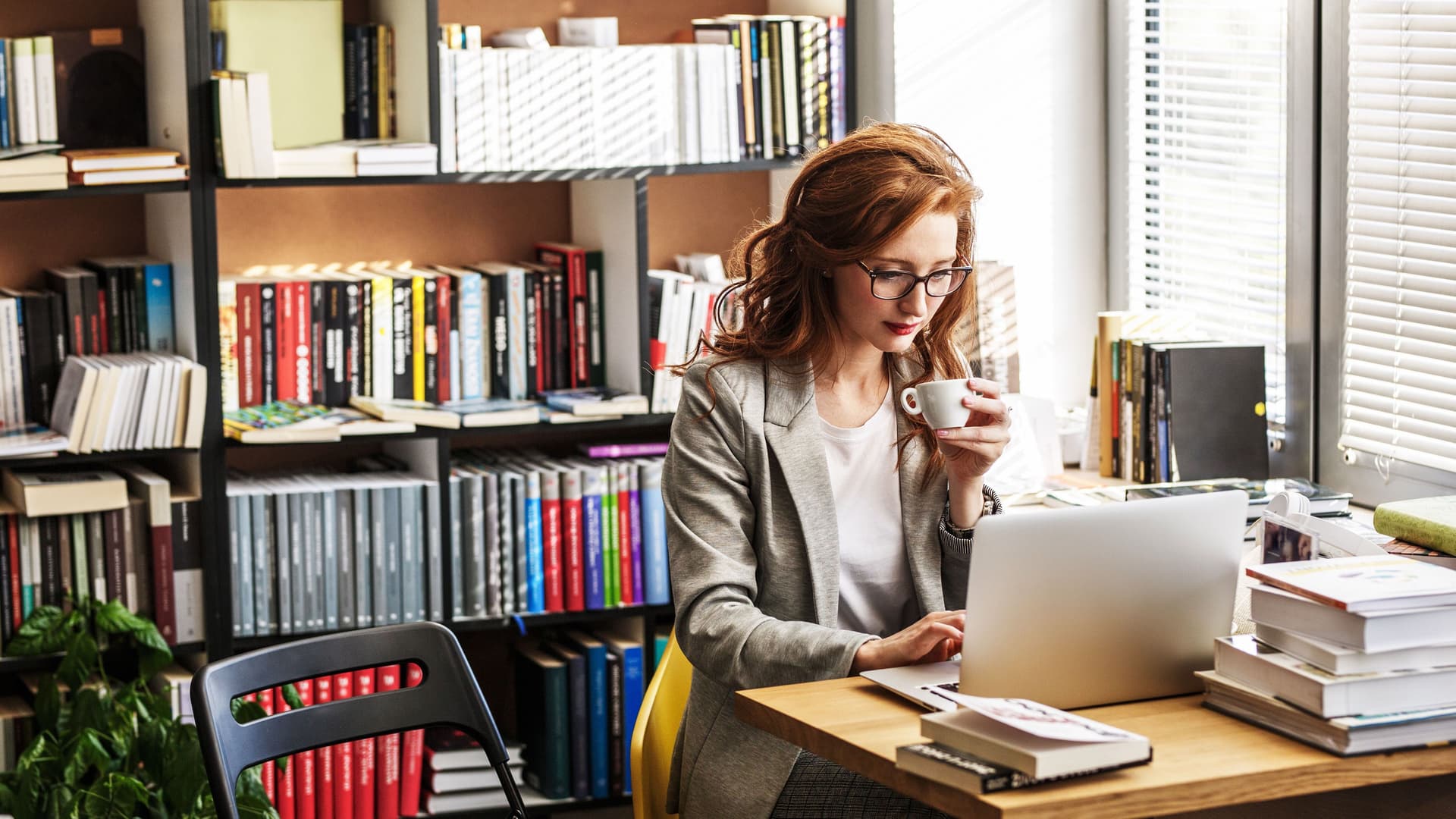 Frau mit Blazer und Brille sitzt vor einem Laptop mit einer Tasse in der Hand. Neben ihr auf dem Tisch und hinter ihr im Regal sind Bücher.