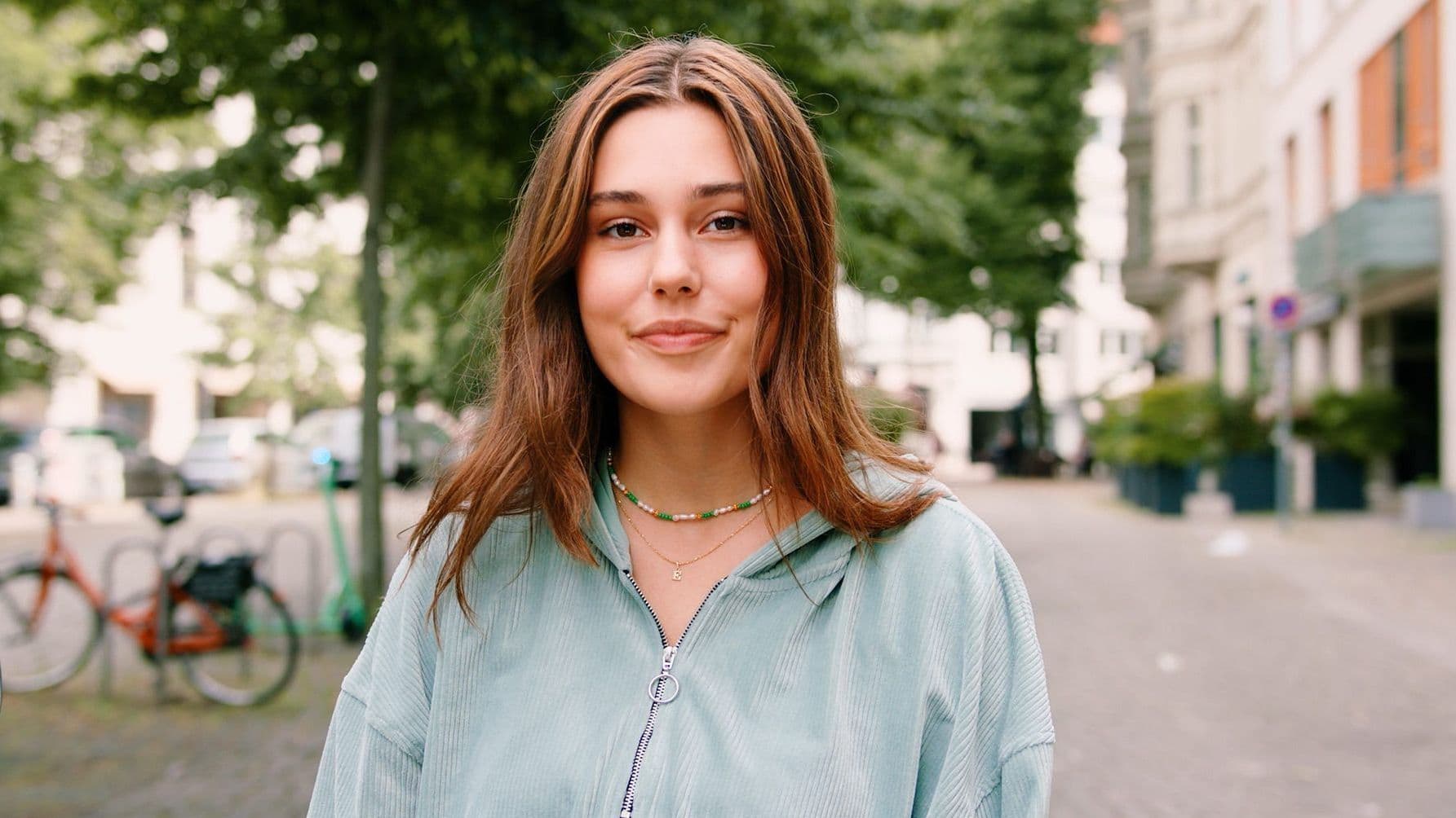 Eine junge Frau steht frontal und mit dem Blick zur Kamera auf einem Bürgersteig. Im Hintergrund sieht man ein urbanes Stadtleben.