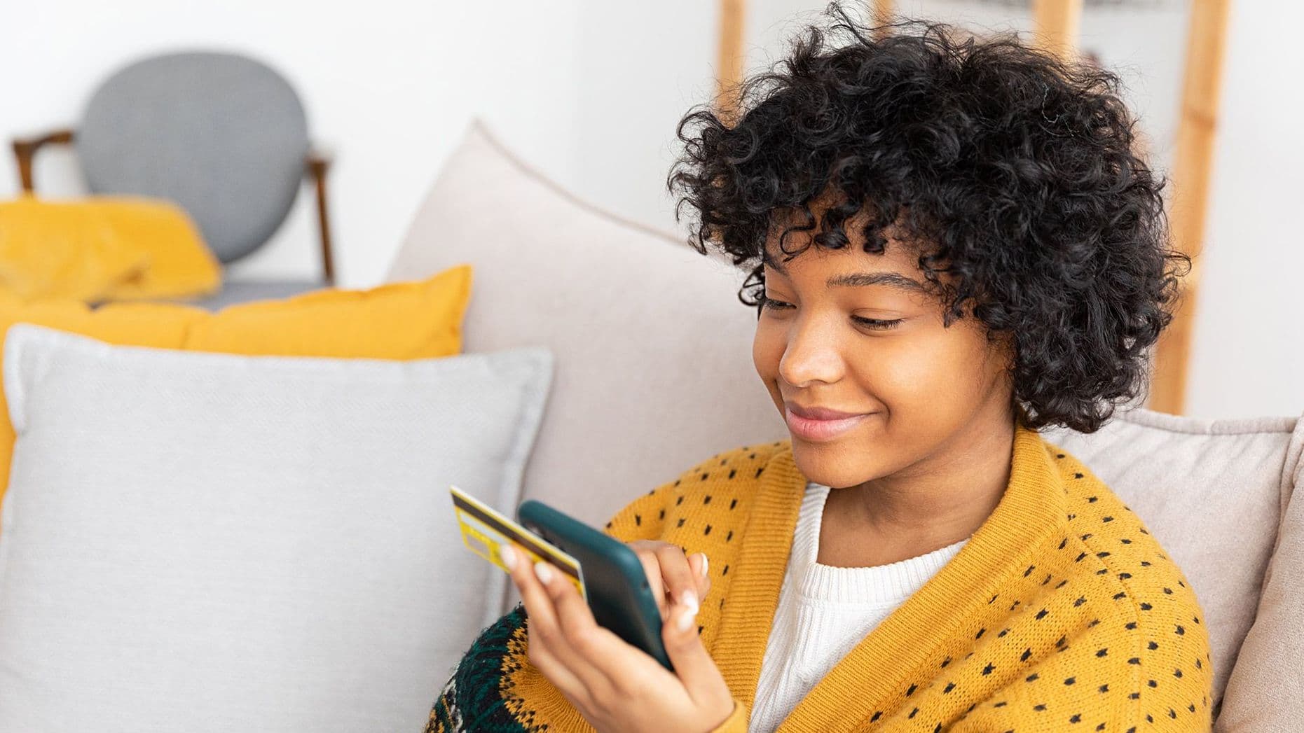 Eine junge Frau sitzt auf einem Sofa und hält neben einer goldenen Kreditkarte, ein Smartphone in den Händen. Sie schaut aufs Display. 