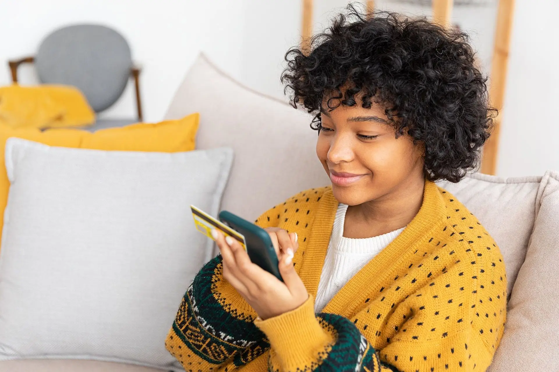 Eine junge Frau sitzt auf einem Sofa und hält neben einer goldenen Kreditkarte, ein Smartphone in den Händen. Sie schaut aufs Display. 
