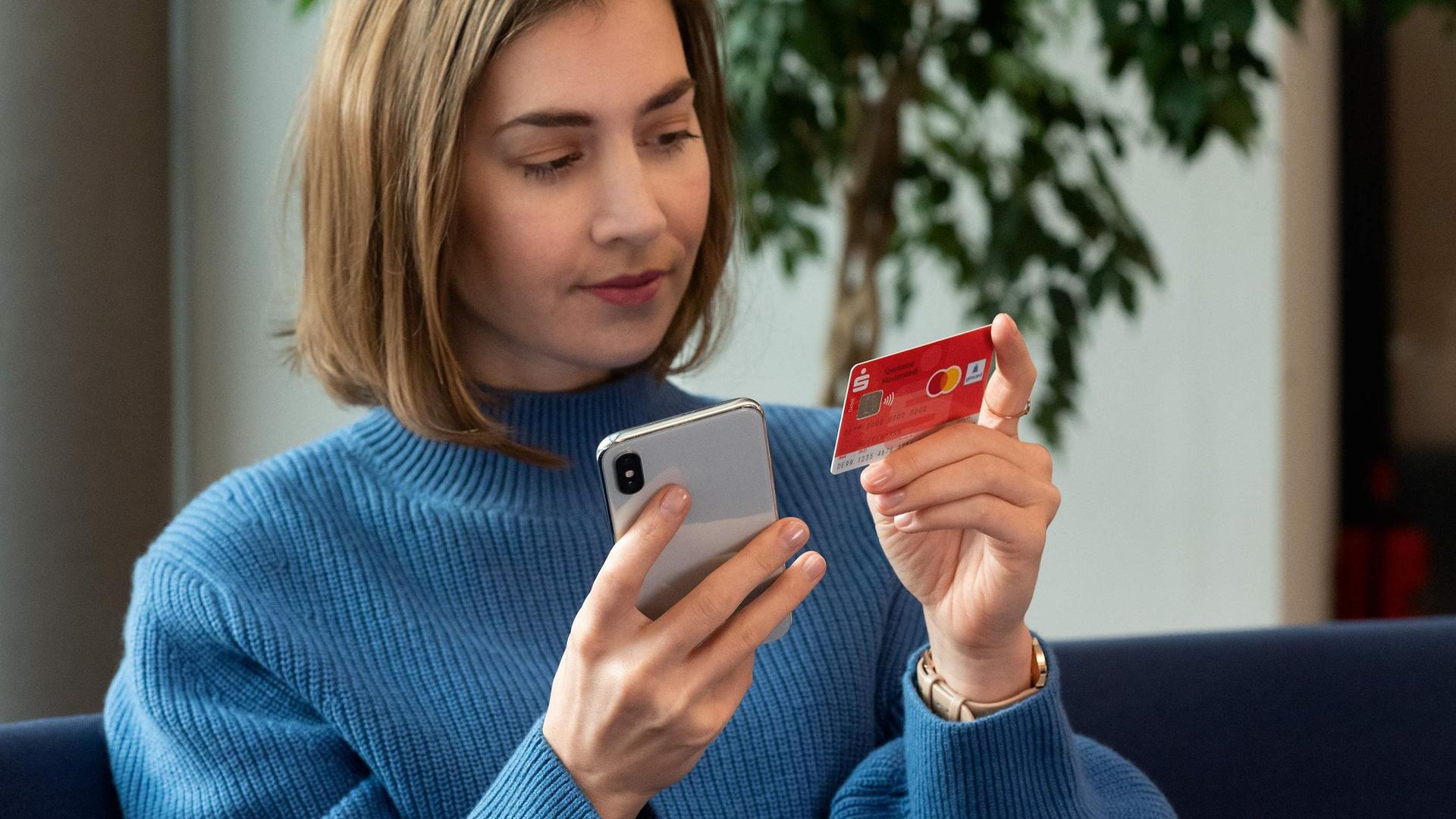 Eine junge Frau sitzt auf einem Sofa und hat ein Handy und ihre Sparkassenbankkarte in jeweils einer Hand. Sie gibt Daten von der Bankkarte in ihr Handy ein.
